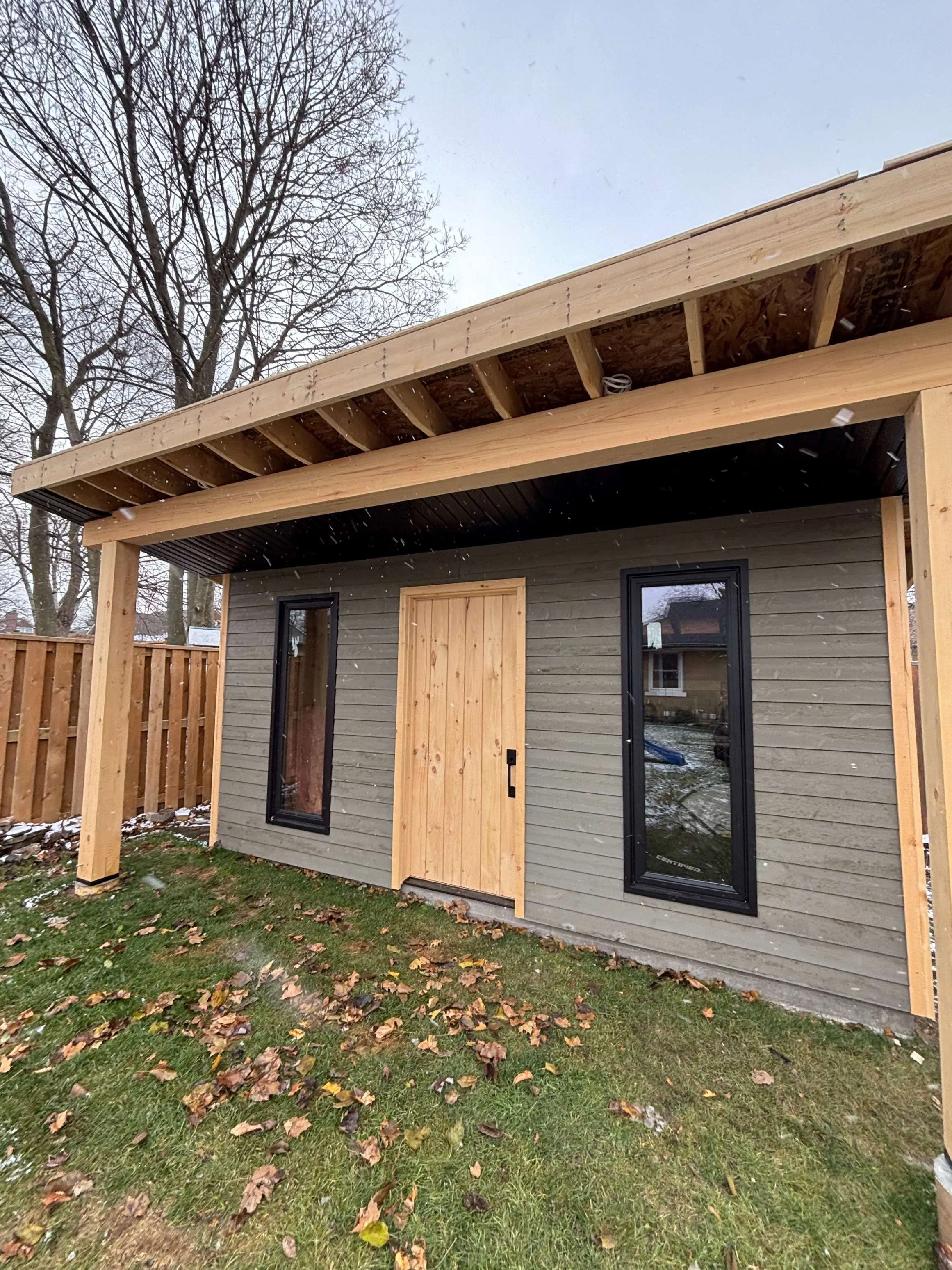 A small house under construction with a wooden frame and partially installed siding, featuring black-framed windows, a wooden door, and a covered porch area, set in a yard with green grass and fallen leaves, with a large tree in the background and light snowfall.
