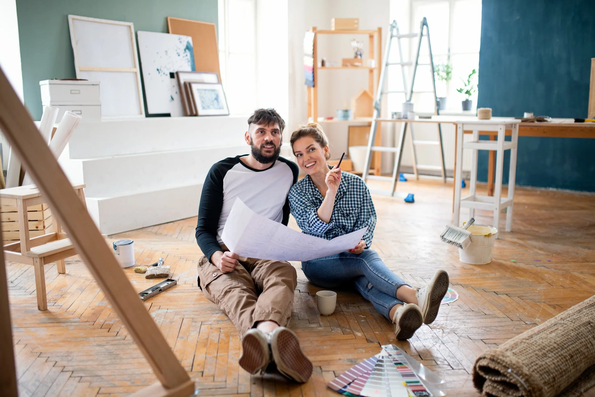 Two people sitting on the floor of an art studio or workshop, looking at a large sheet of paper or blueprint. The studio has art supplies, paint swatches, frames, and ladders around, with a large window in the background.
