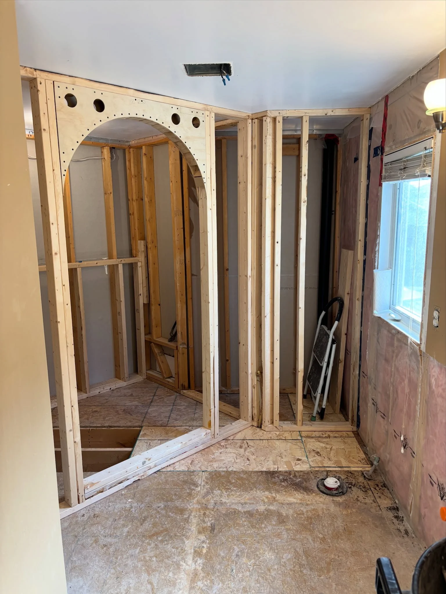 Interior room under construction with wooden framing, exposed wiring, a small window, and construction tools on the floor.