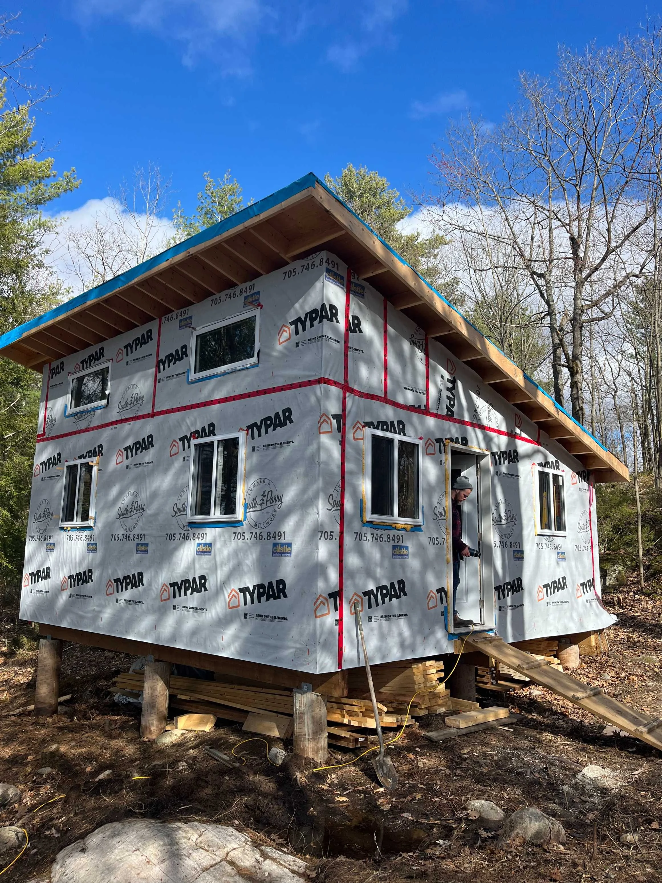 A two-story house under construction in a wooded area, covered with Typar building wrap, with multiple windows installed. The roof is partially complete with wooden support beams. Construction tools and materials are scattered around the site.
