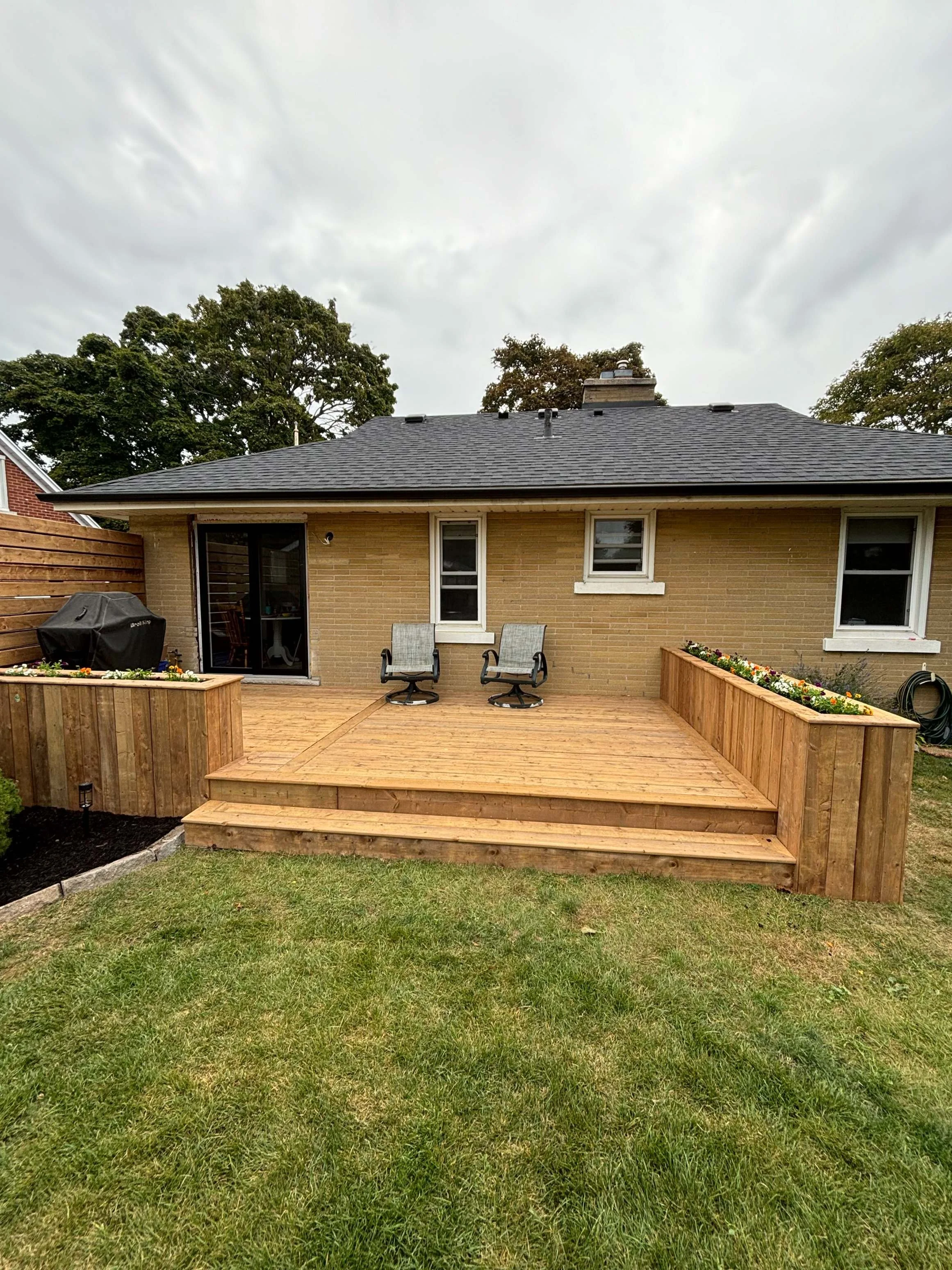 A backyard patio with a wooden deck, two chairs, flower beds, and a house with a brick exterior and sliding glass door. There are trees and a cloudy sky in the background.