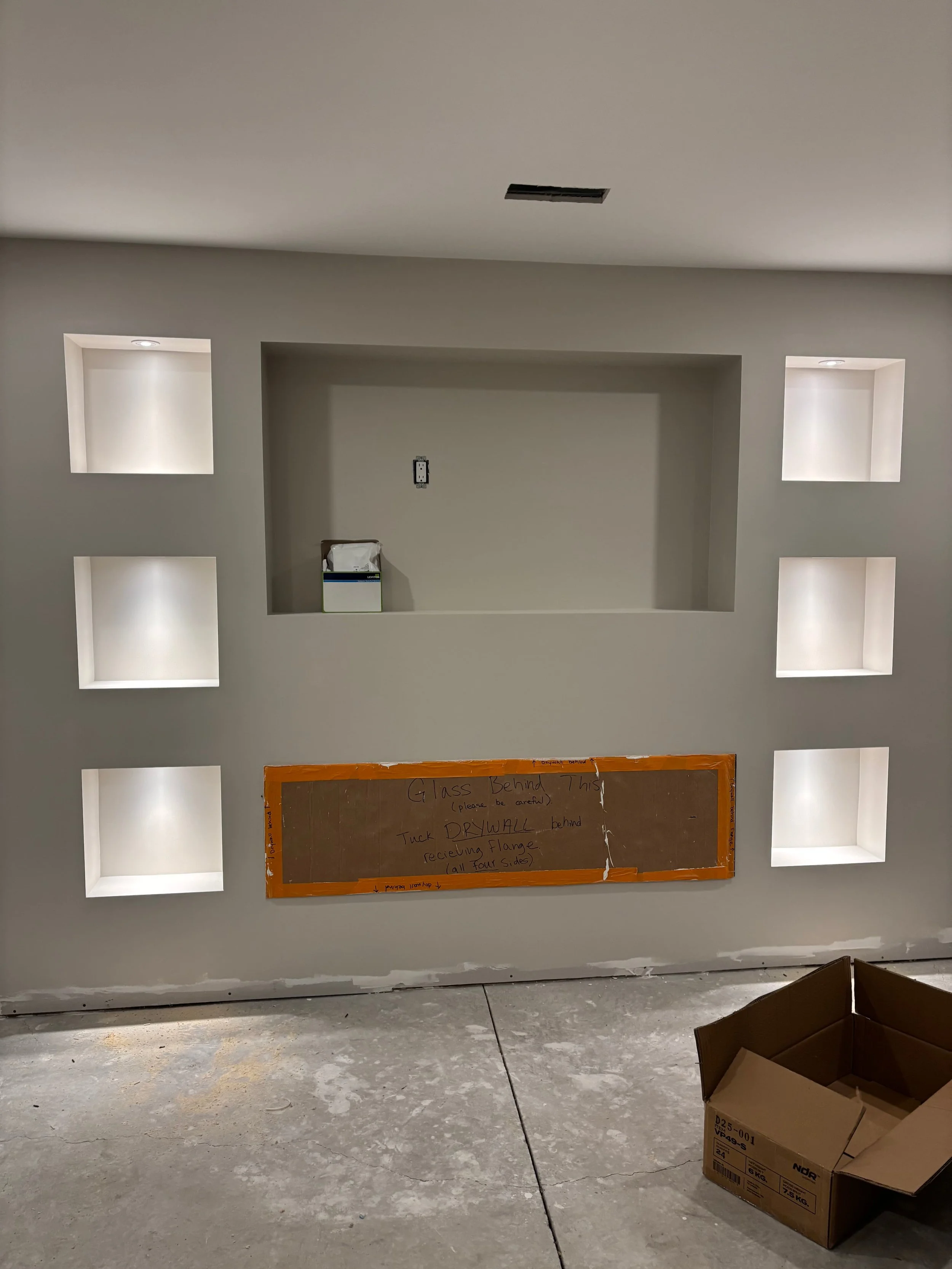 Construction site with a wall featuring built-in square shelves lit from above, a bulletin board with handwritten instructions, and an open cardboard box on the floor.