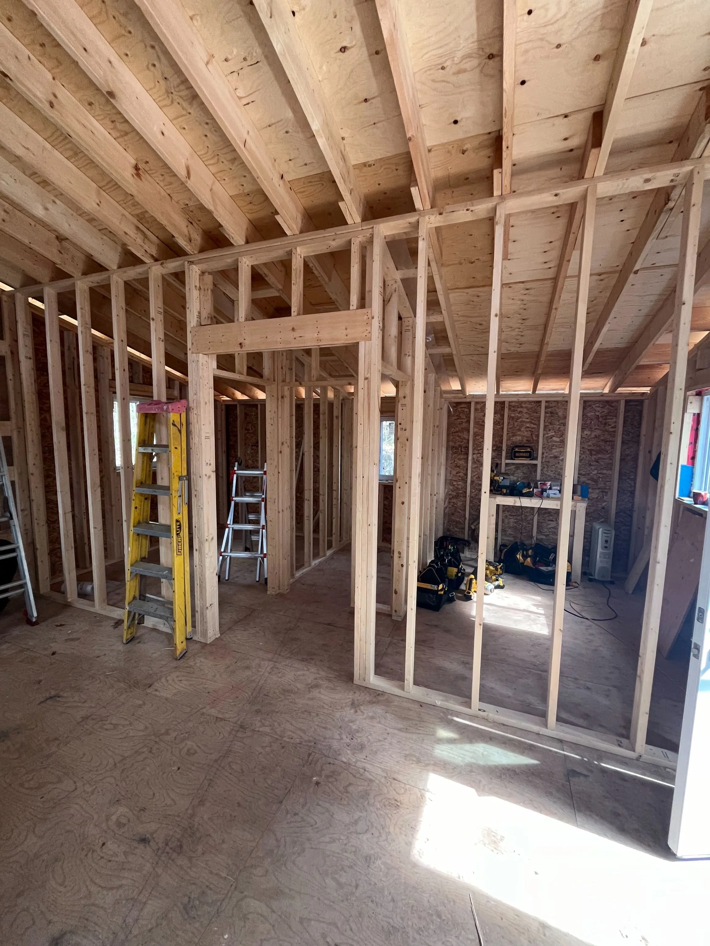 Interior view of a house under construction with exposed wooden framing, ladders, and construction tools on the floor.