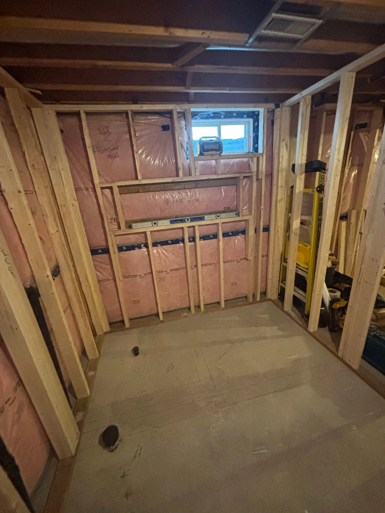 Interior view of a partially constructed room with wooden framing, pink insulation, small window, and construction tools.