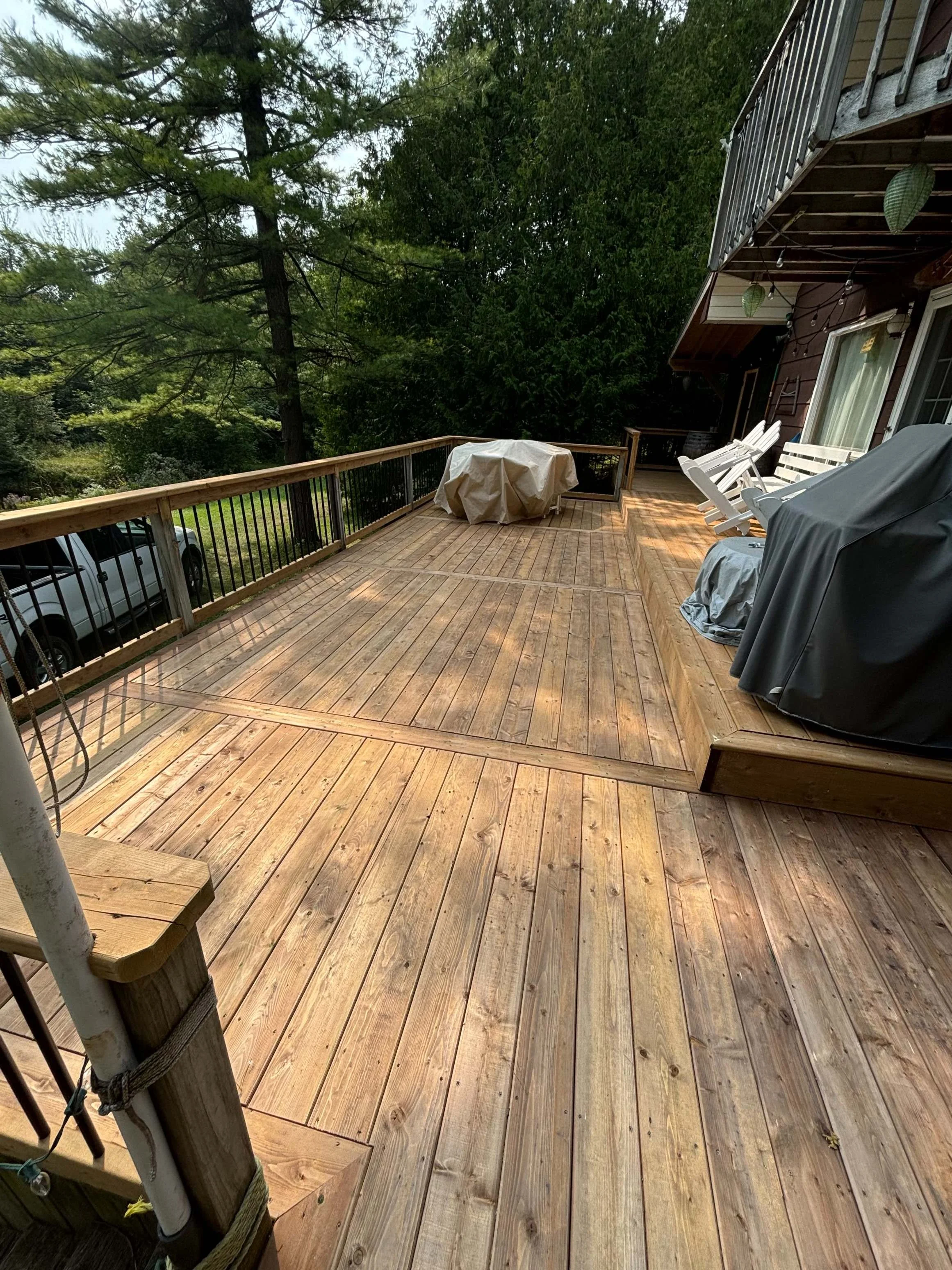 A spacious outdoor wooden deck with several white chairs, a covered barbecue grill, a round table with a cover, and a balcony with hanging plants, overlooking a green, tree-filled landscape.