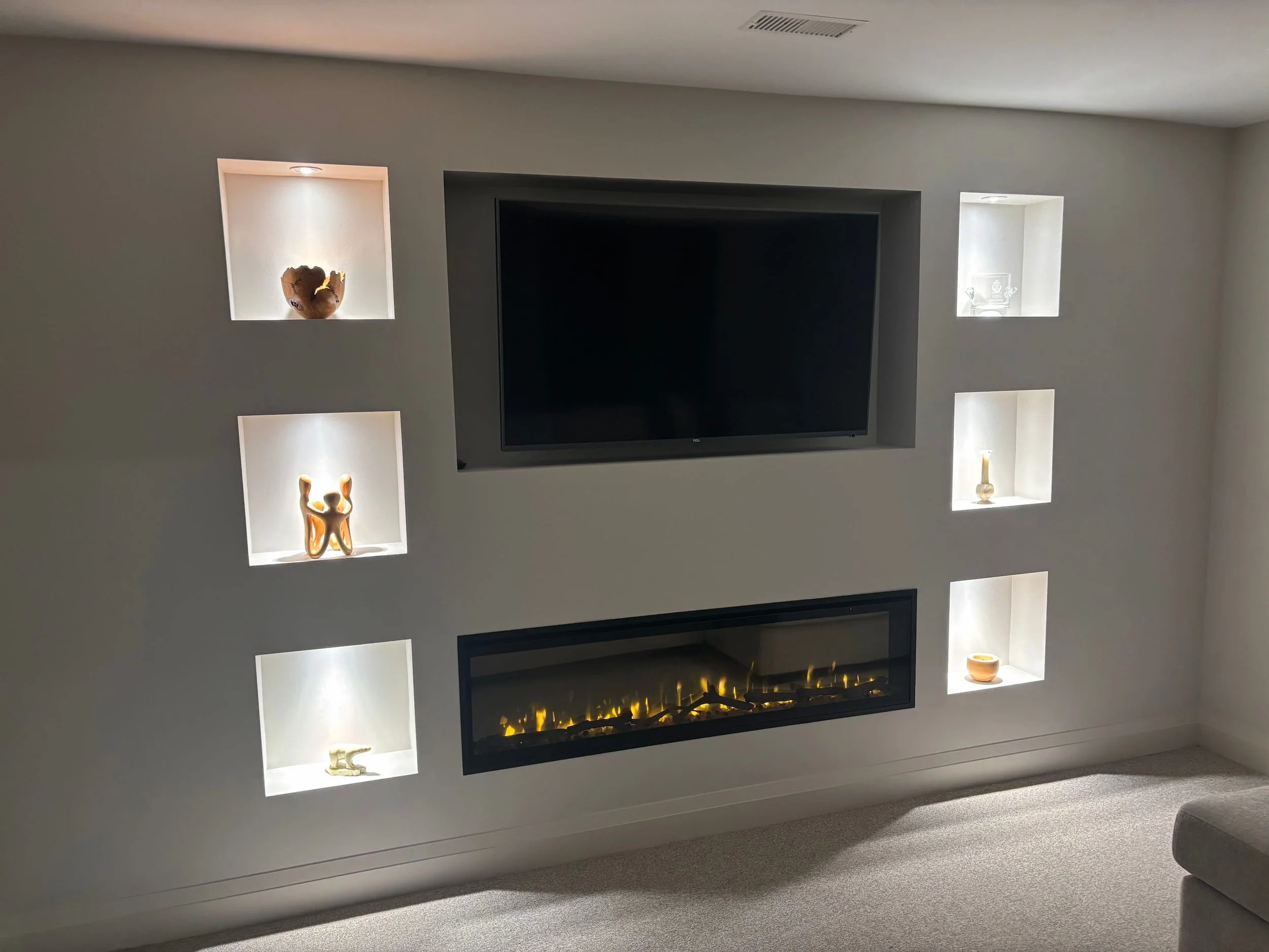 Modern living room with built-in illuminated display shelves, a flat-screen TV, and a fireplace below the TV. The shelves showcase decorative items including sculptures and vases.