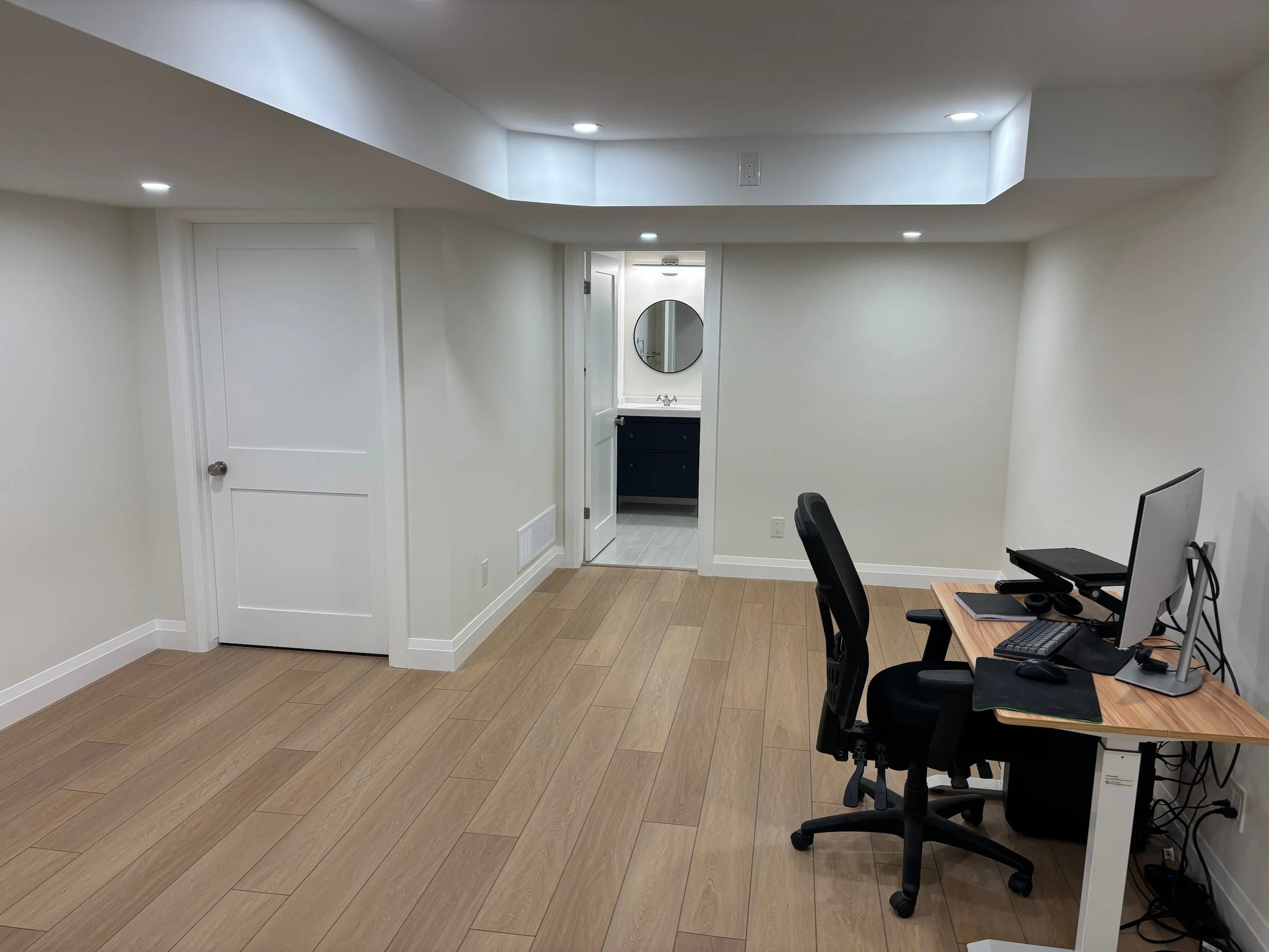 A small room with beige walls, wood flooring, and a white ceiling. There is an open doorway leading to a small bathroom with a mirror, sink, and dark-colored vanity. The room has a desk with computers, monitors, and office supplies, and a black offic