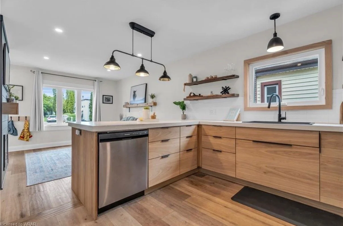 Modern kitchen with wooden cabinets, stainless steel dishwasher, black faucet, white countertop, and two windows with views of greenery outside; hanging pendant lights above the counter, and floating wooden shelves with decorative items.