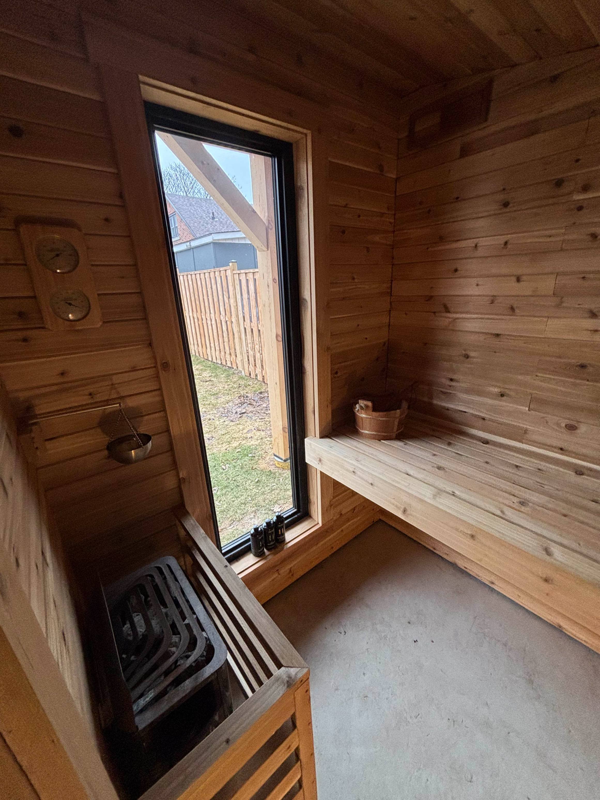 Inside a small wooden sauna room with a window showing outside a fence and some houses. The room has a wooden bench and a bucket, with a heater on the floor and some bottles on the windowsill.