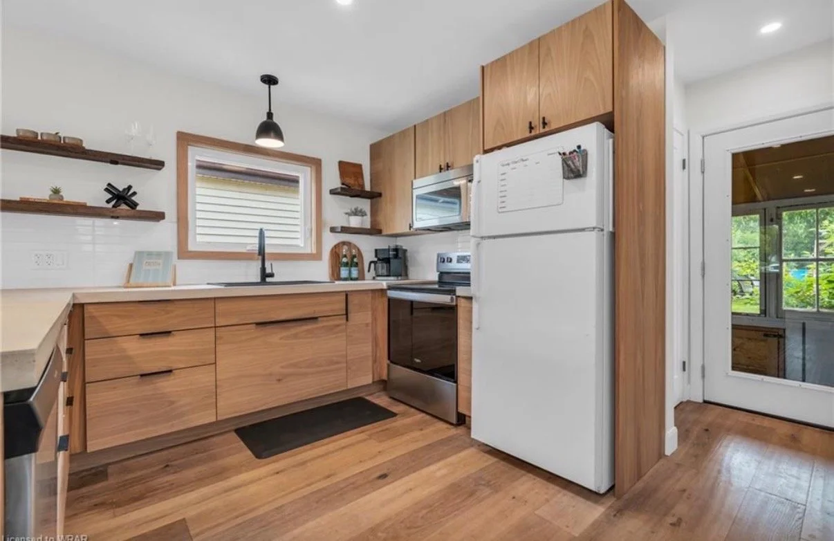 Kitchen with wooden cabinets, a white refrigerator, black oven, microwave, and a small window above the sink, decorated with floating shelves and a small potted plant.