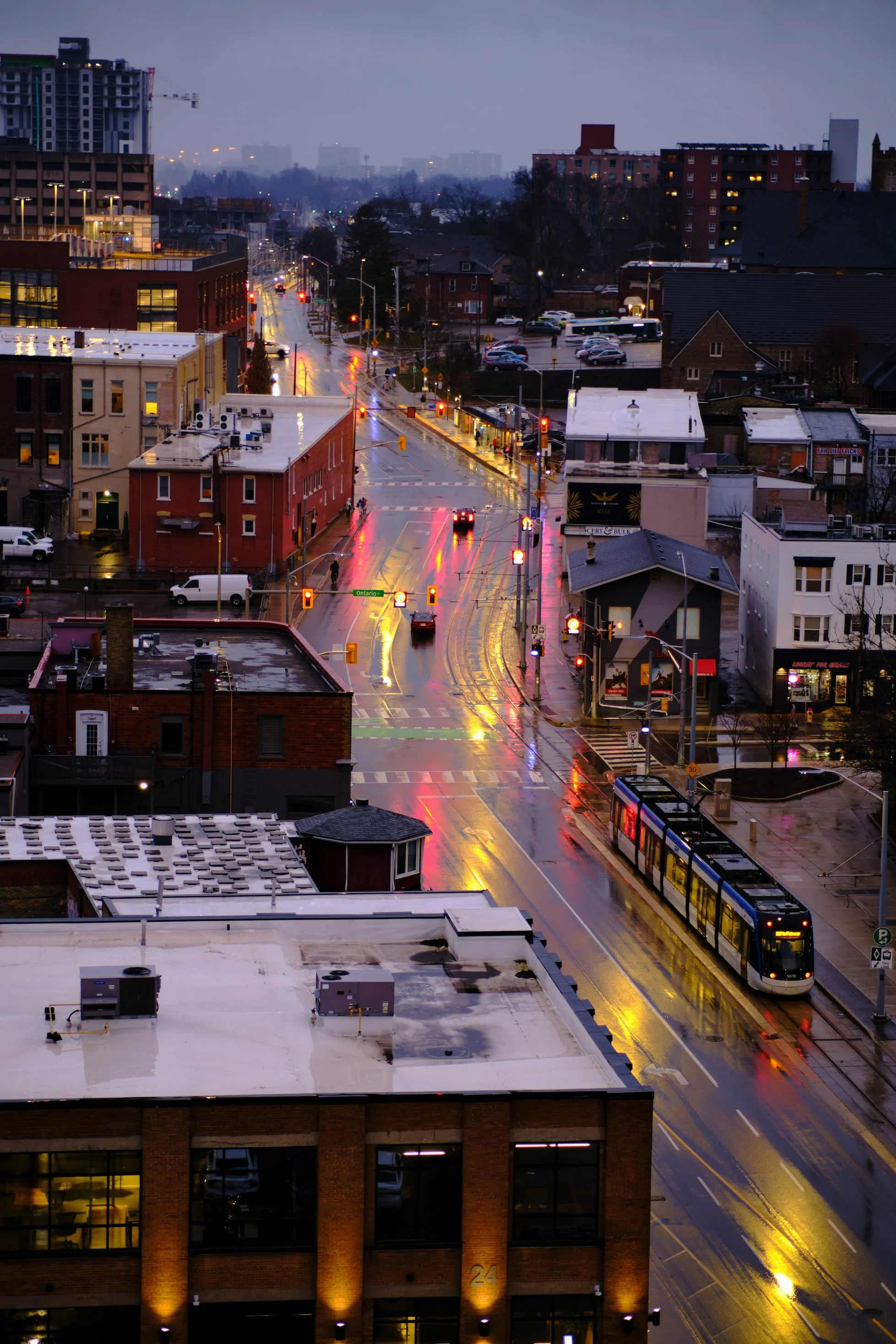 City street during rainy evening with reflections on wet pavement, buildings with illuminated windows, and a train at the station.