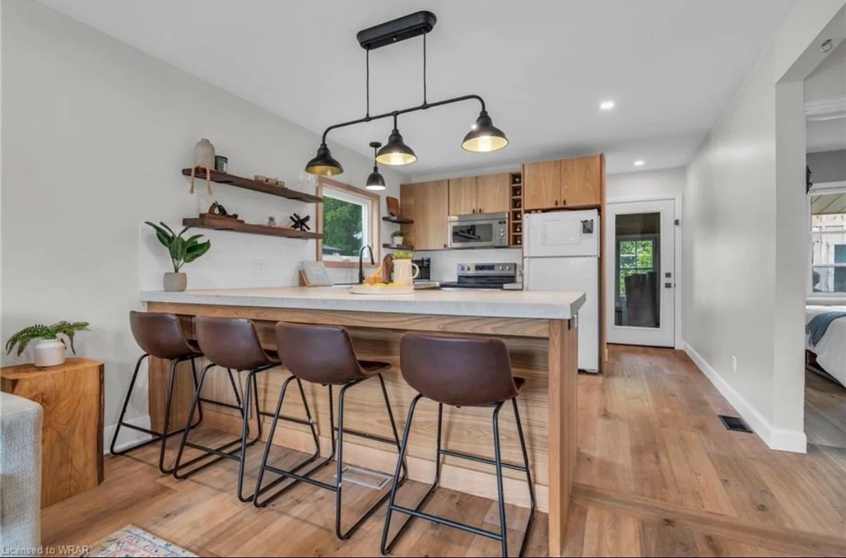 Modern kitchen with wood accents, a white countertop island with four brown leather bar stools, black pendant lights, wooden cabinets, a window above the sink, and a door leading outside.