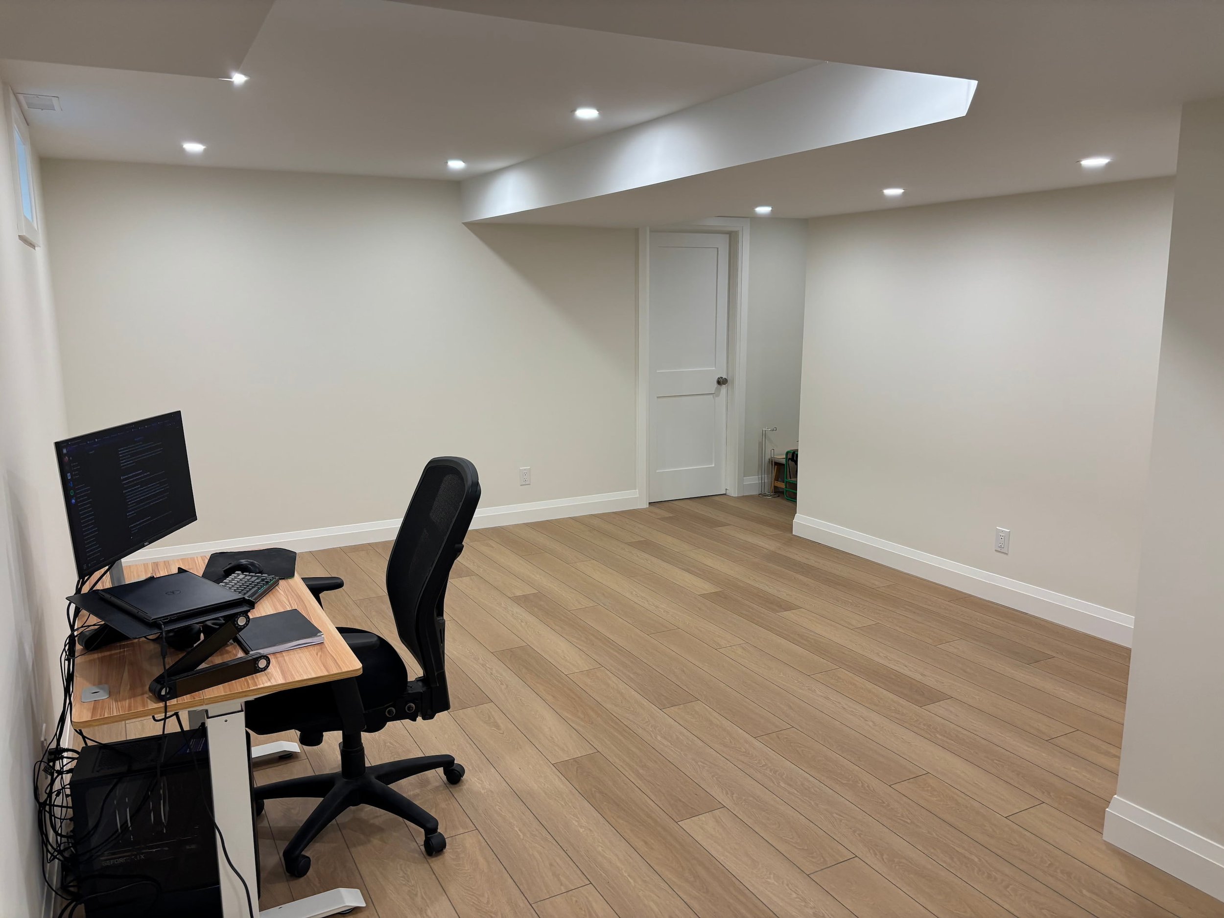 An office space with white walls and ceiling, hardwood flooring, a white door, a desk with a computer, and a black office chair.