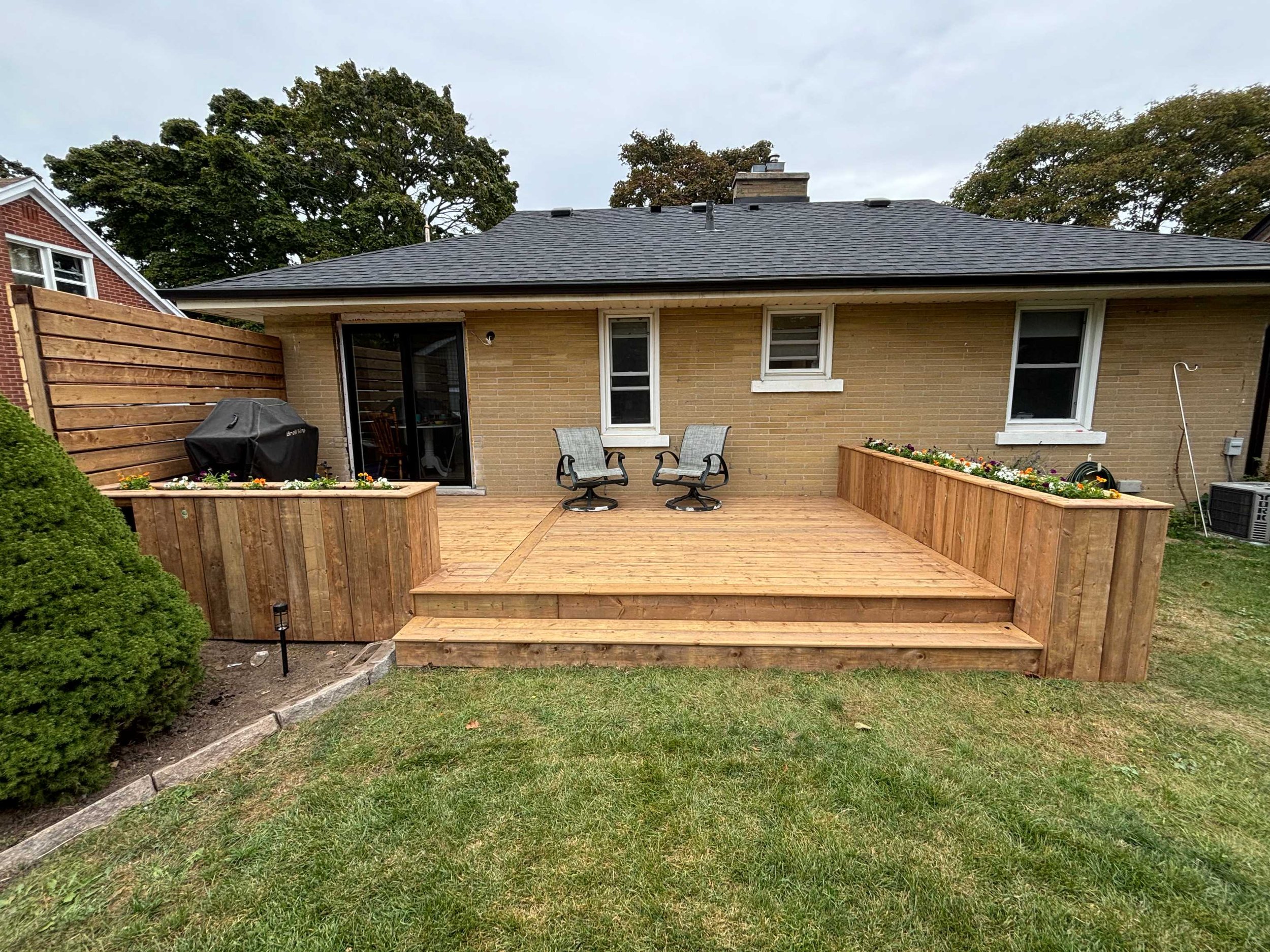Backyard patio with wooden deck, two chairs, a grill, and flower beds, with a house in the background and trees in the yard.
