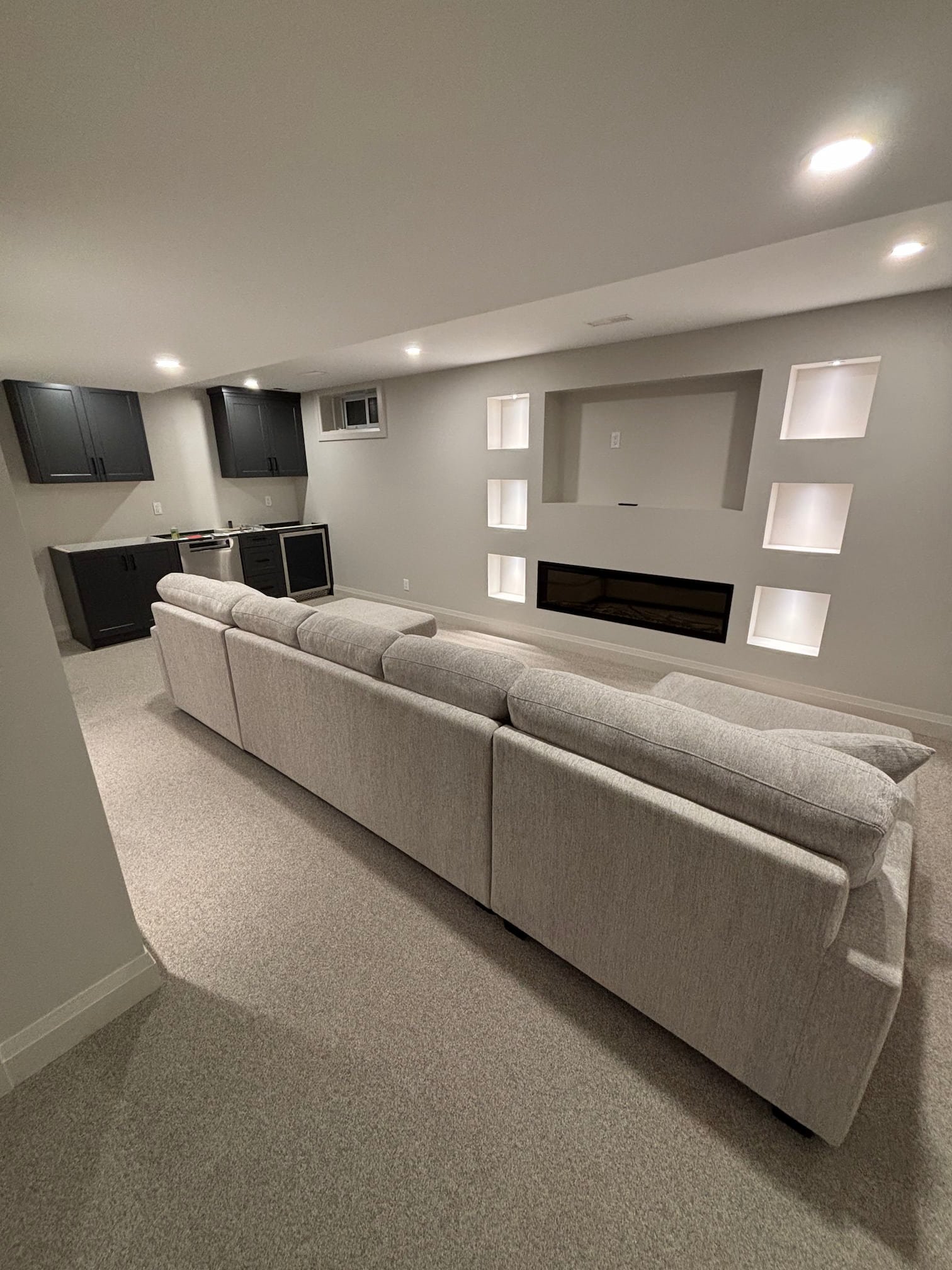 A cozy basement living room with a gray sectional sofa, built-in wall fireplace, black kitchen cabinets, and lighting from recessed ceiling lights.