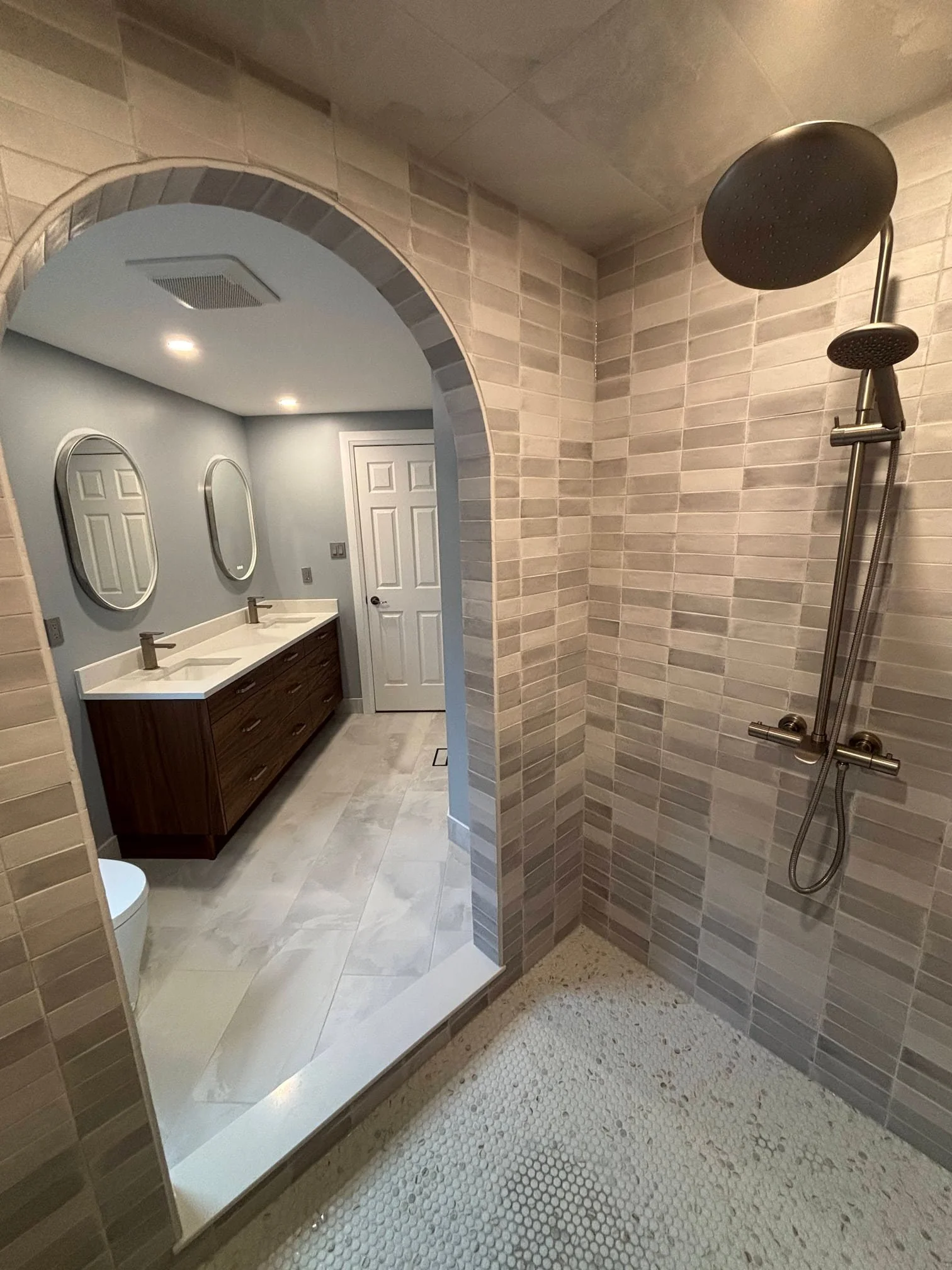 A modern bathroom with a shower area featuring beige tiles, a rainfall shower head, a double vanity with a wooden cabinet, and two oval mirrors on a blue wall.