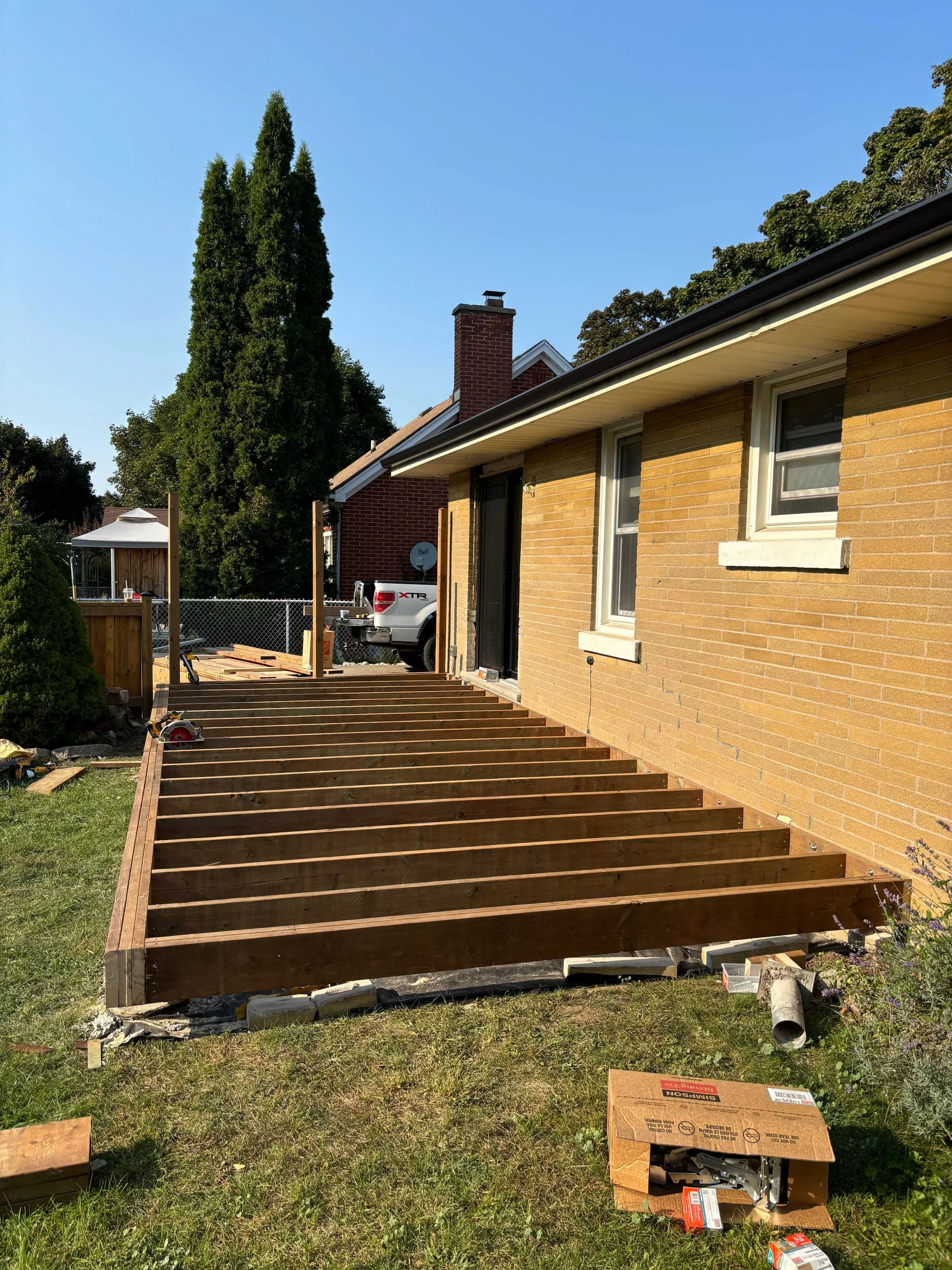 Backyard construction site with a partially built wooden deck next to a yellow brick house, a pickup truck, and a tall evergreen tree under a clear blue sky.