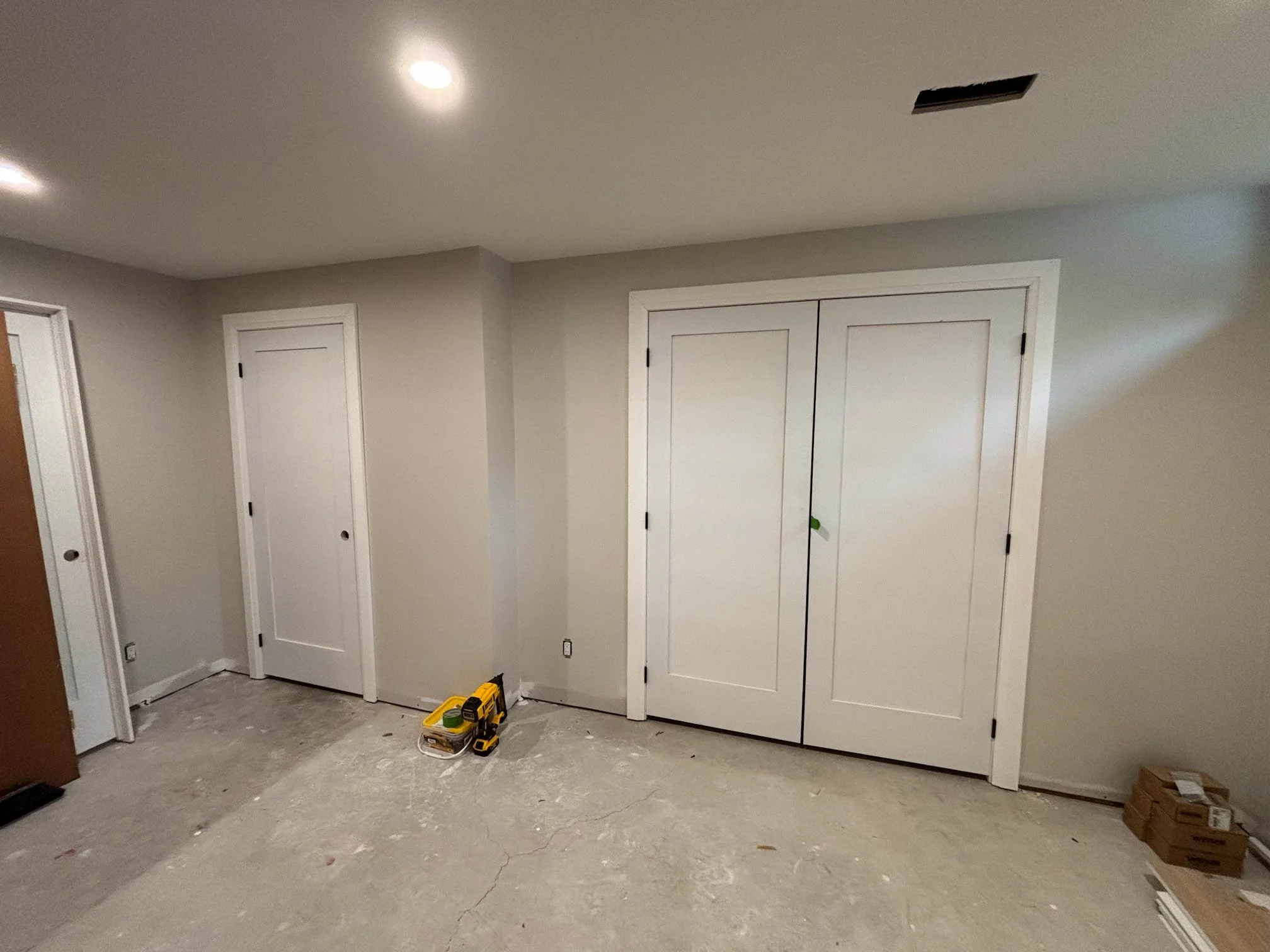 The room appears to be under construction or renovation with a concrete floor, some wooden boxes, and tools scattered around. There are three white closet doors on the wall and a partially visible door on the left. The ceiling has two recessed lights