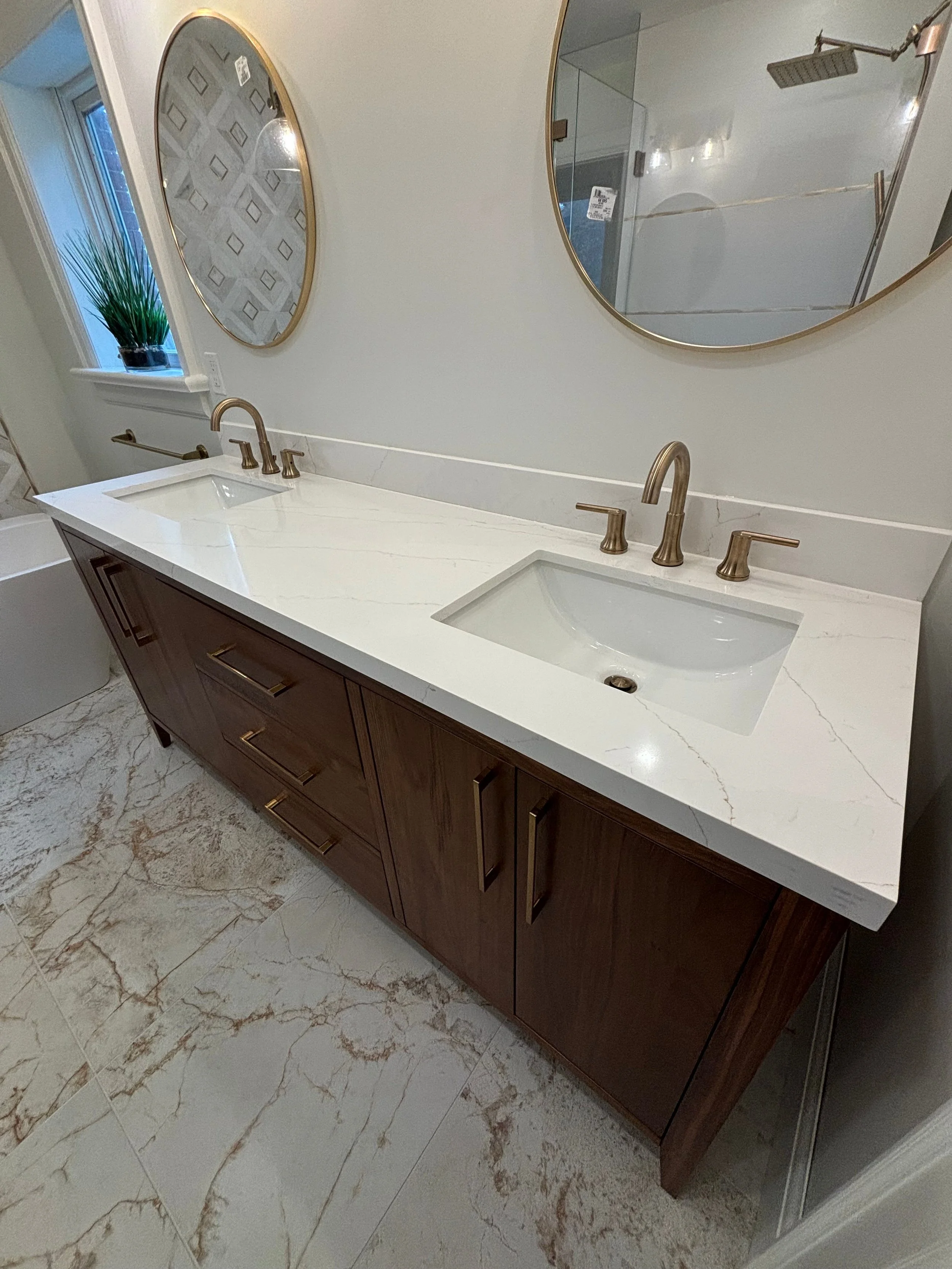 A double vanity bathroom sink with a white marble countertop, two brushed gold faucets, and a dark wood cabinet. Two round mirrors with gold frames are mounted above the sinks. A window with a plant on the windowsill is visible in the background, and a walk-in shower with a glass enclosure and a rain showerhead is seen on the right side. The bathroom floor is made of light-colored marble tiles with brown veining.