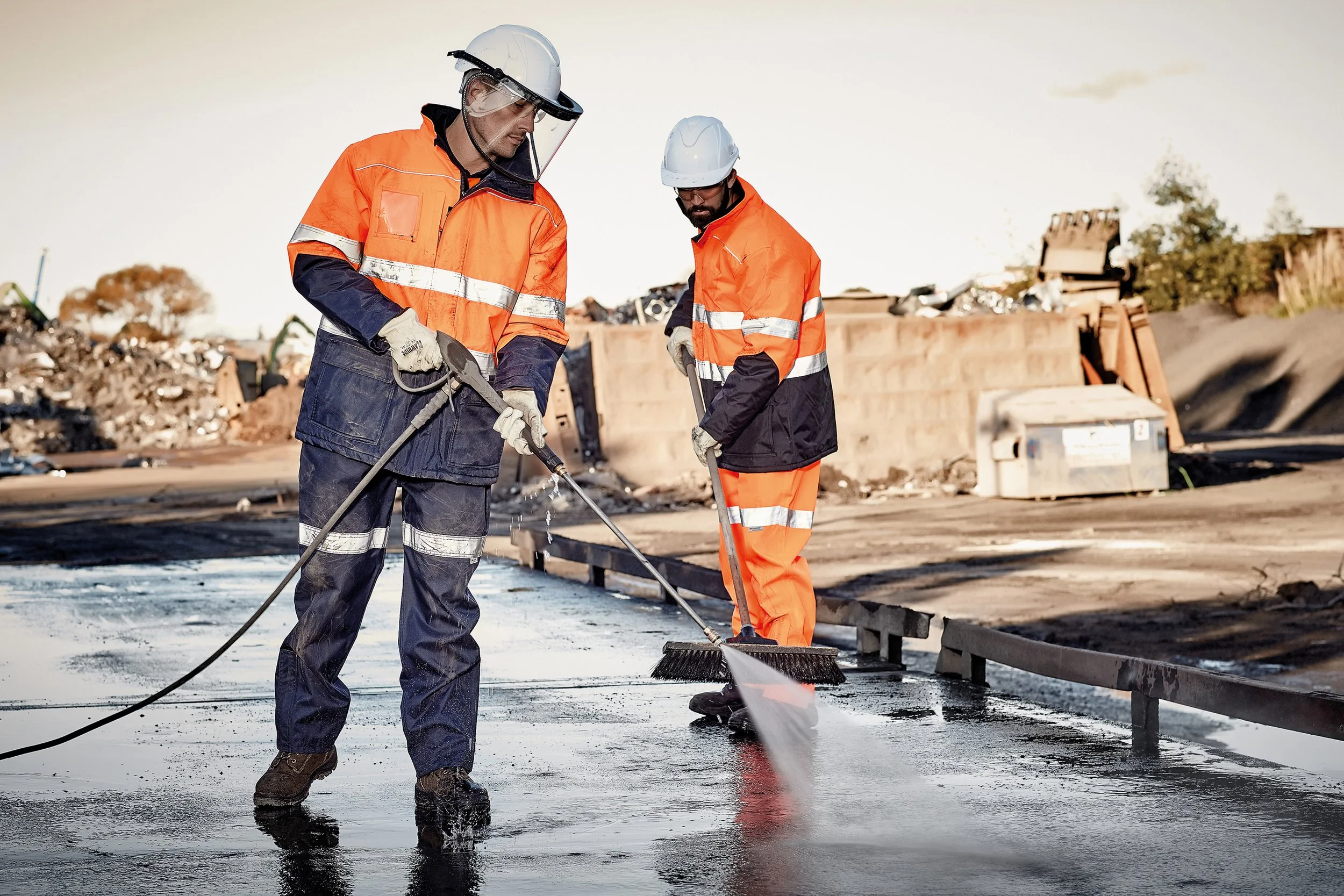 Three people standing outdoors near water on a cloudy day, with sailboats in the background. Two men and one woman, with one man wearing an orange jacket, one man in an orange shirt with reflective stripes, and the woman in a navy jacket, looking ahead.
