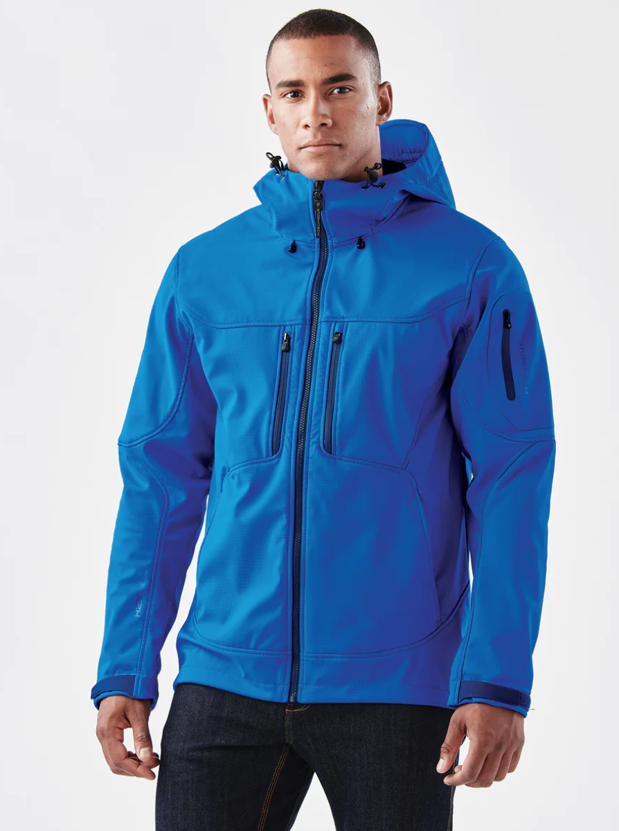 A man leaning against a wooden wall outdoors, wearing a white button-up shirt with two front pockets, with a blurred green landscape in the background.