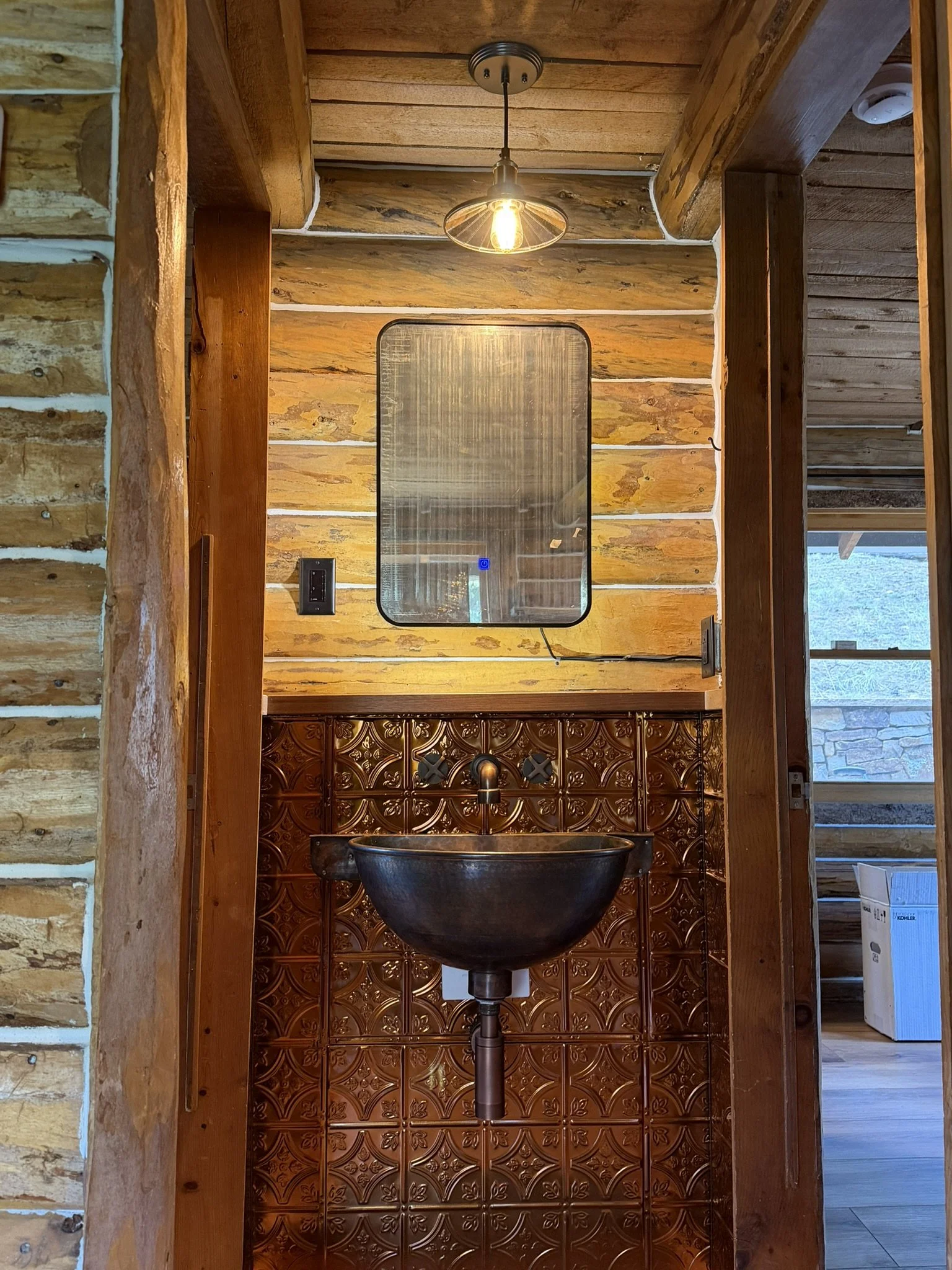 A rustic bathroom with wooden paneling, a mirror, and a black vessel sink with a bronze faucet. There is a hanging light bulb overhead and a window to the right showing outdoors. The walls feature decorative brown tiles with floral patterns.
