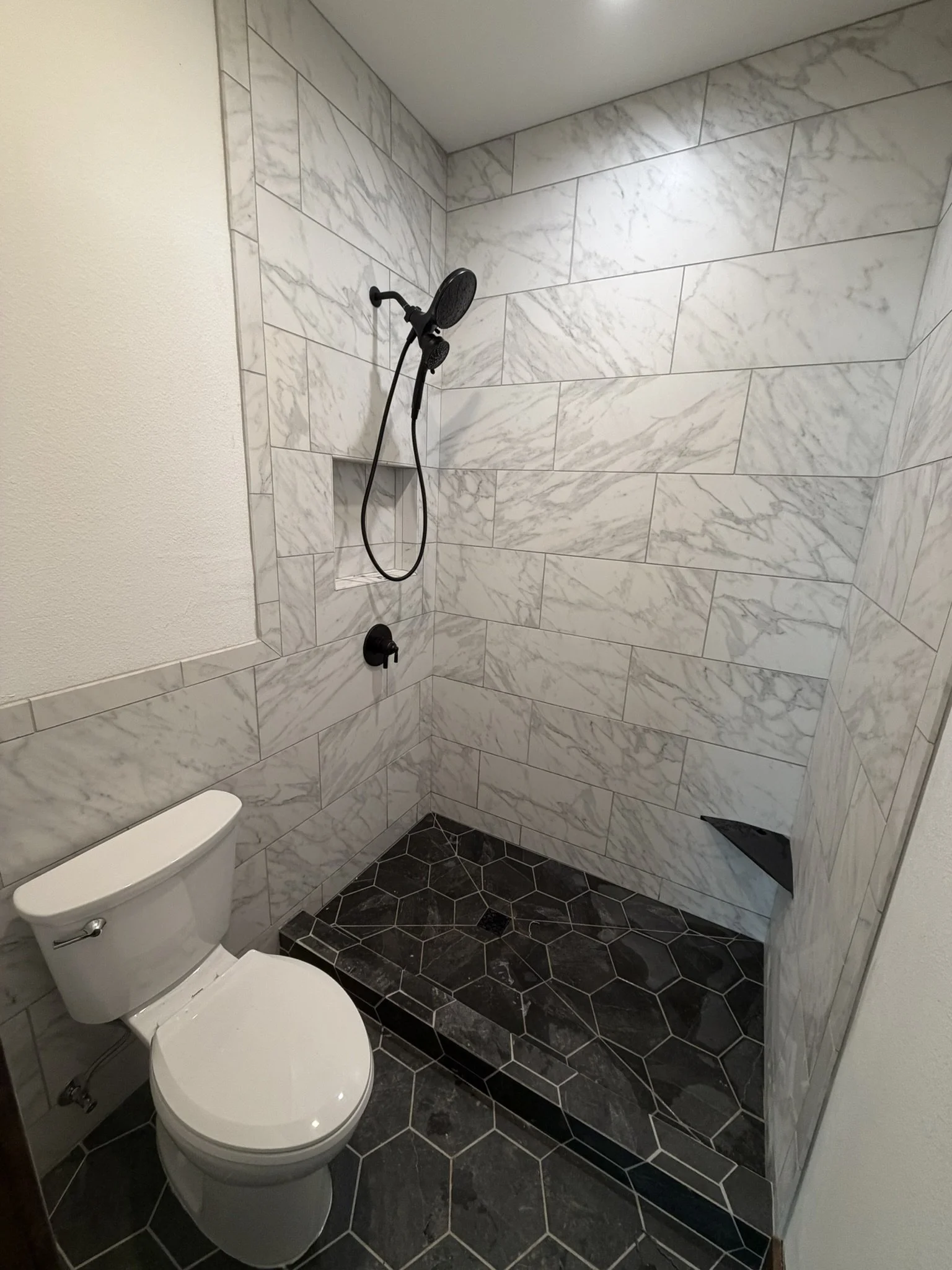 A small bathroom with white marble walls, a toilet, and a walk-in shower with black handheld showerheads. The floor has black hexagonal tiles with white grout.