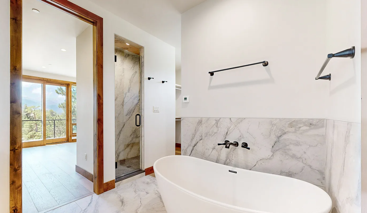 Modern bathroom with a freestanding bathtub, marble tiles, a walk-in shower, and a view of a bright room with large windows and a balcony.