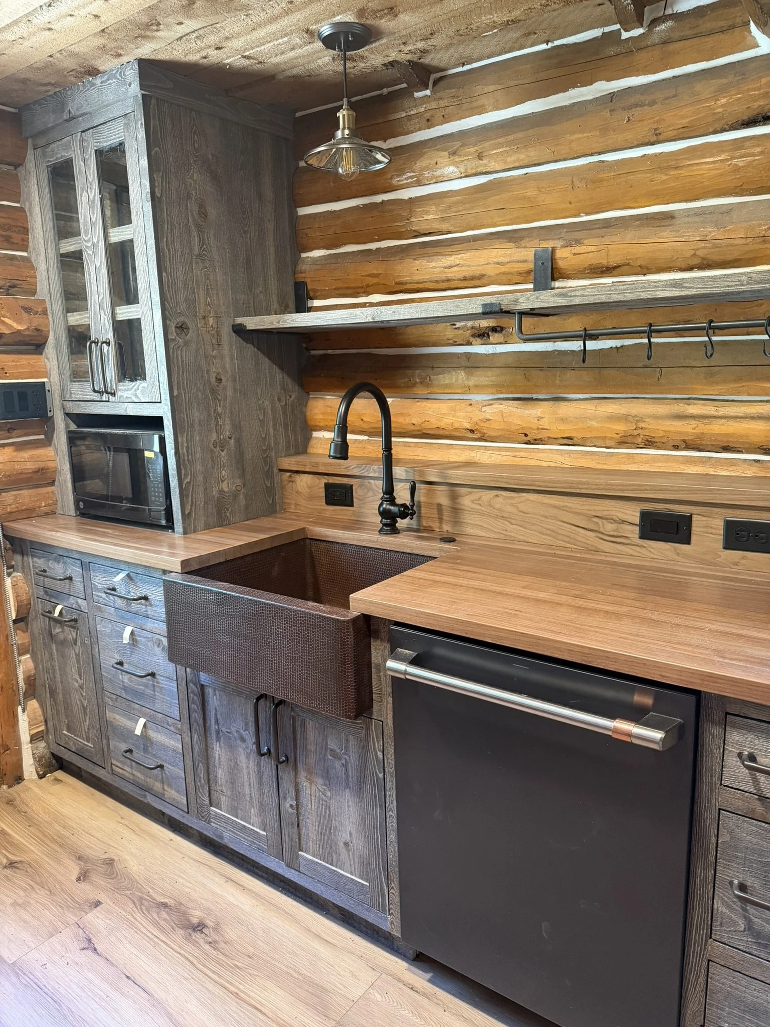 Rustic kitchen with wooden cabinets, a copper sink, black faucet, and black appliances, set against a wood-paneled wall with open shelving and a hanging light fixture.