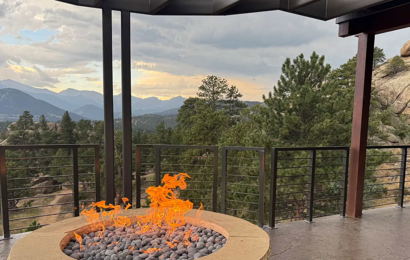 A balcony with a fire pit filled with gray stones and flickering flames, overlooking a mountainous landscape with pine trees under a partly cloudy sky.