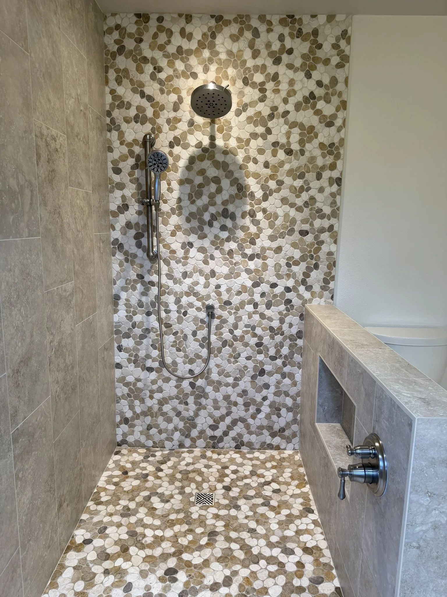 A walk-in shower featuring a pebble tile wall and floor, with a round showerhead and a handheld shower attachment, and a small window with a ledge on the right side.