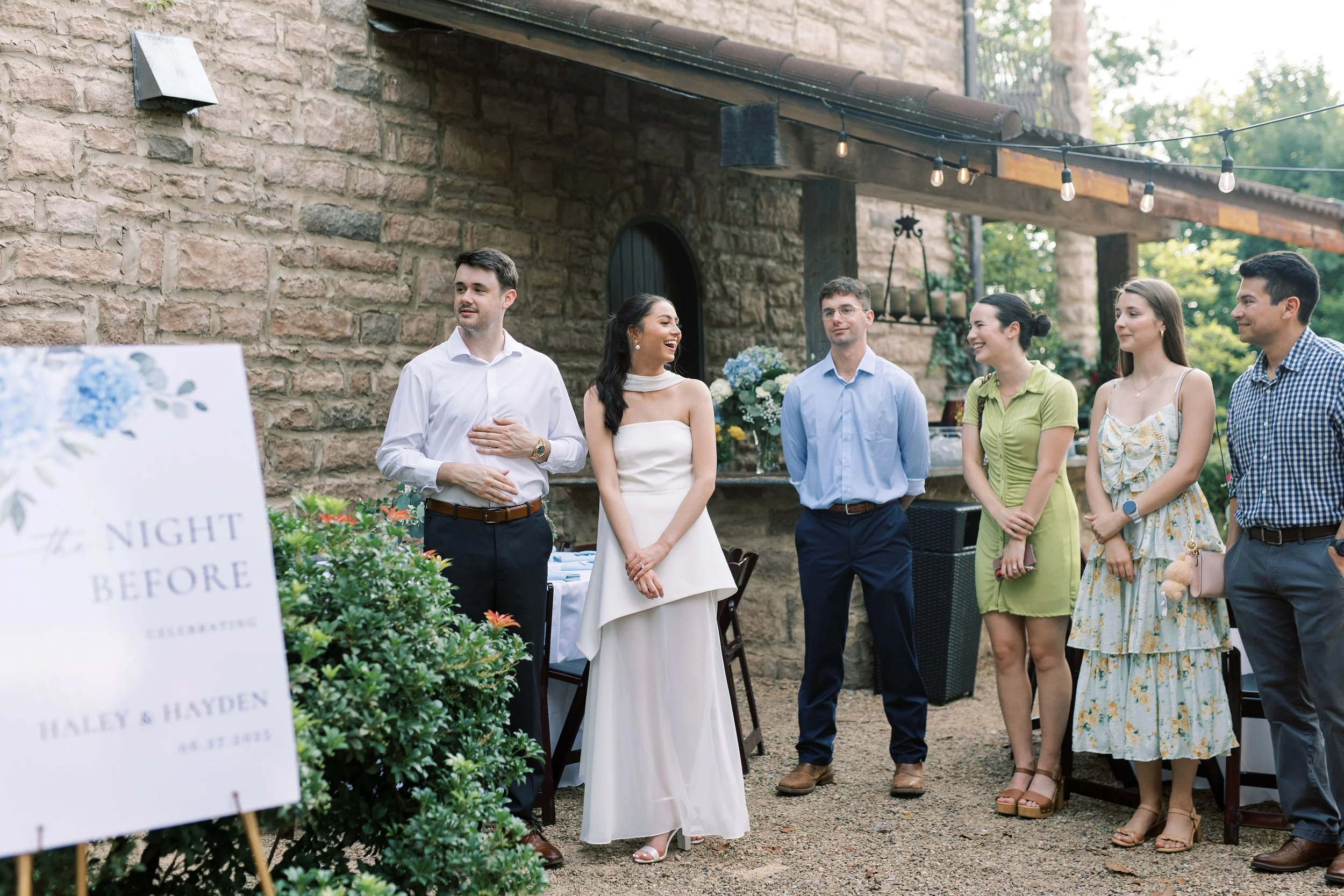 Group of people at a wedding celebration outdoors, with the bride in a white dress in the center, smiling. The groom and friends stand nearby, some with hands on their chest or in pockets. There is a sign in the foreground that reads 'Night Before Celebrations, Haley & Hayden, 04.27.2025' and decorative flowers and string lights in the background.