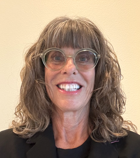 A woman with dark blonde hair in a messy updo, wearing a pink shirt, standing against a beige wall.