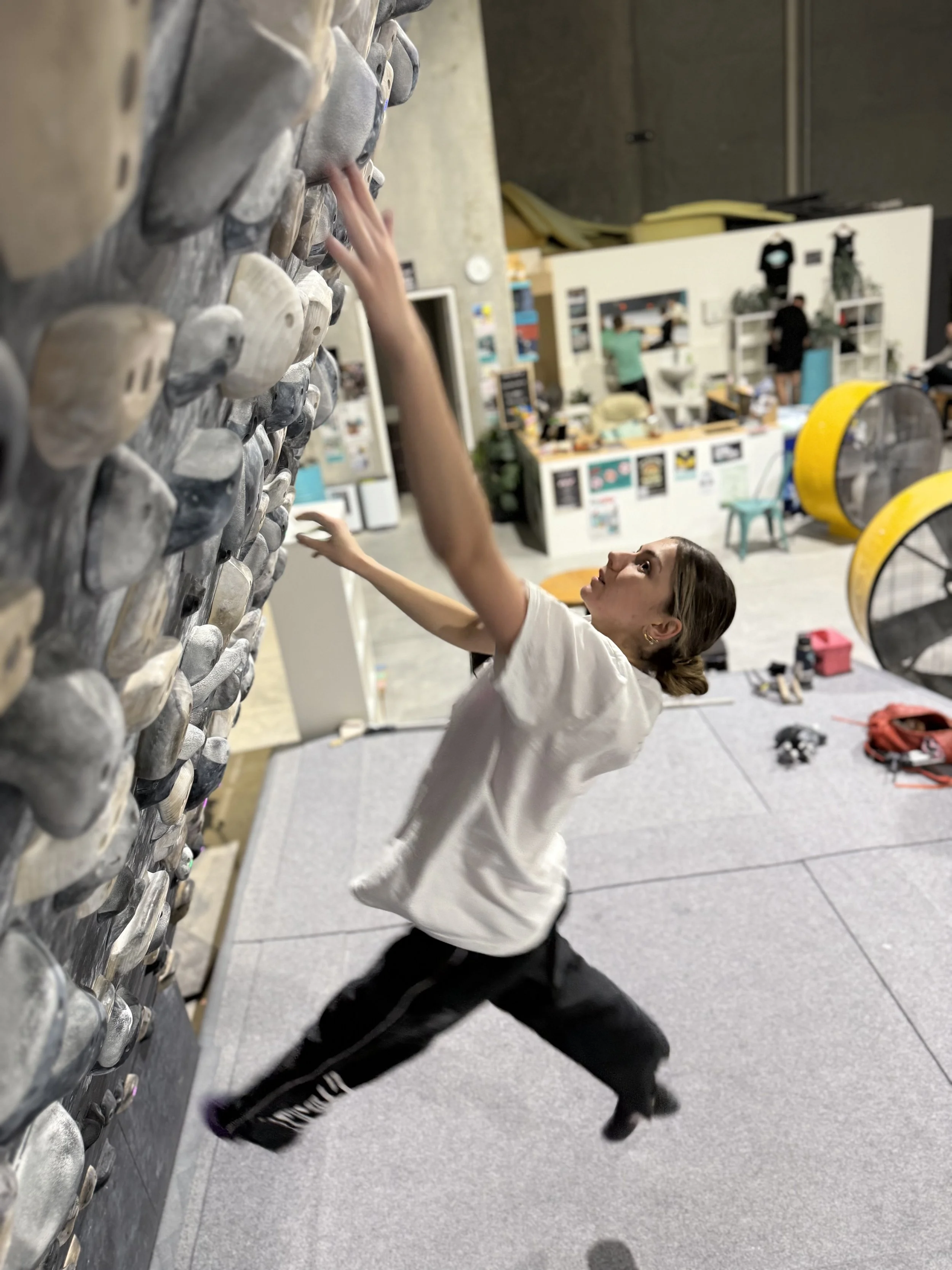 A woman climbing an indoor rock climbing wall with gray handholds, reaching upward with her right hand. She is wearing a white shirt and black pants, and her hair is tied back. The background features a fitness or climbing gym with various equipment and people.