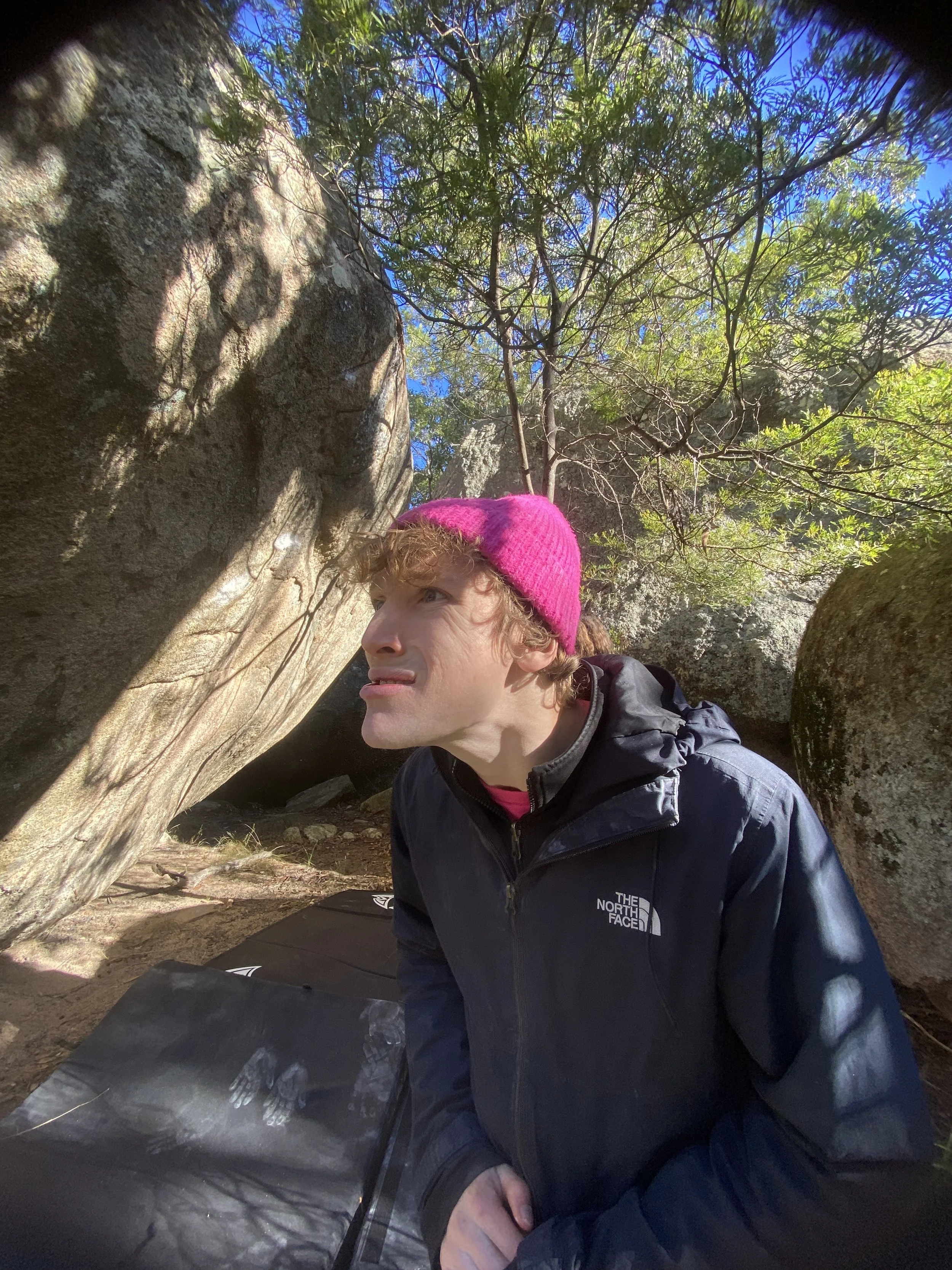 Person with curly hair wearing a pink beanie and black North Face jacket, crouching near large rocks and trees outdoors during daylight.