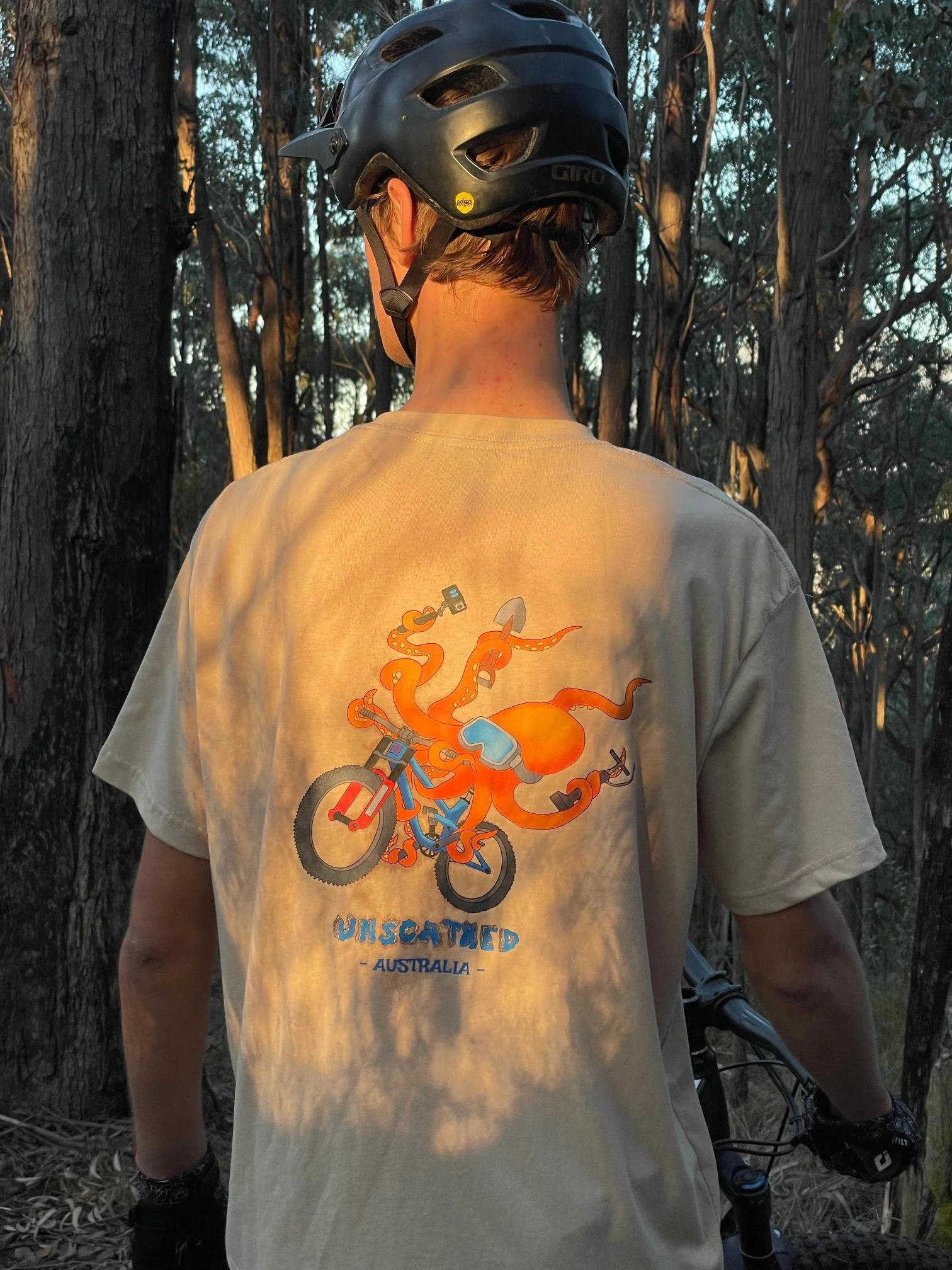 Back of person wearing a beige T-shirt with an illustration of an orange octopus riding a bicycle, with the words 'Unscathed Australia' printed on it, standing in a forested area with trees and sunlight filtering through