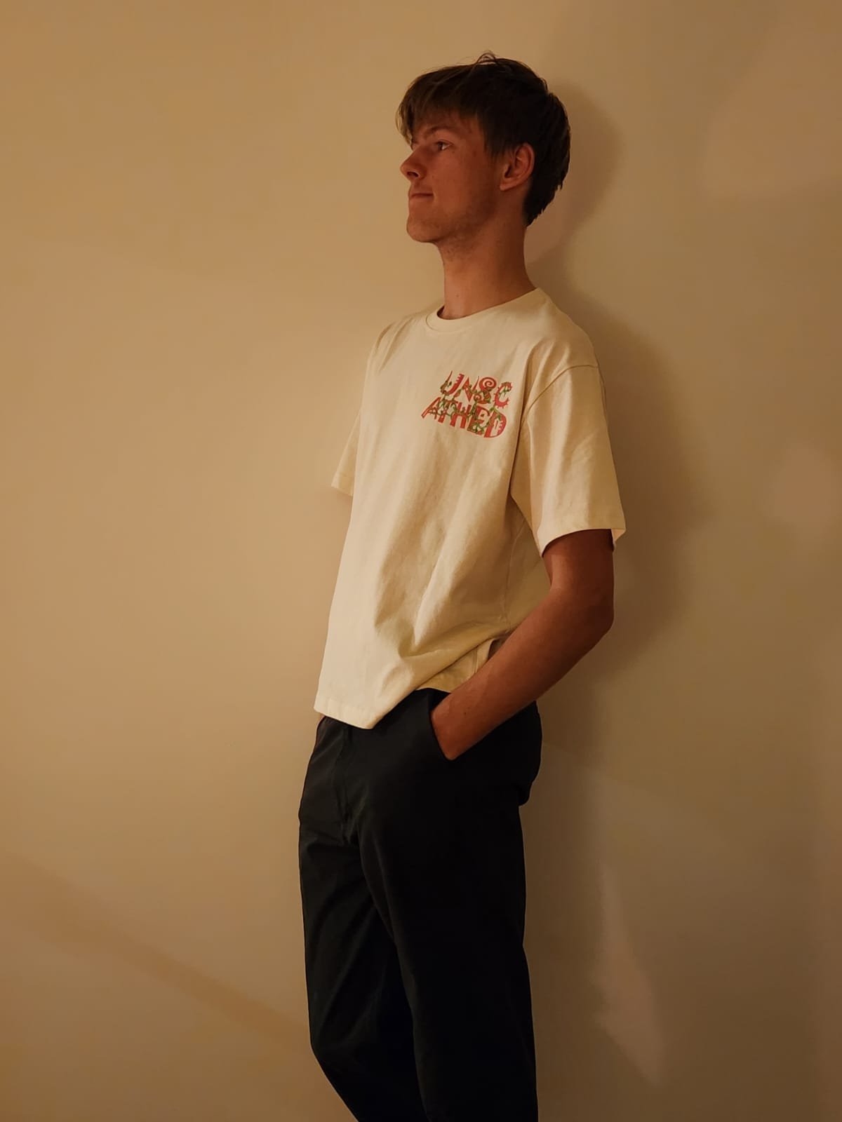 A young man with brown hair standing against a beige wall, wearing a cream t-shirt with red text on the chest and black pants, with his hands in his pockets, looking to the left.