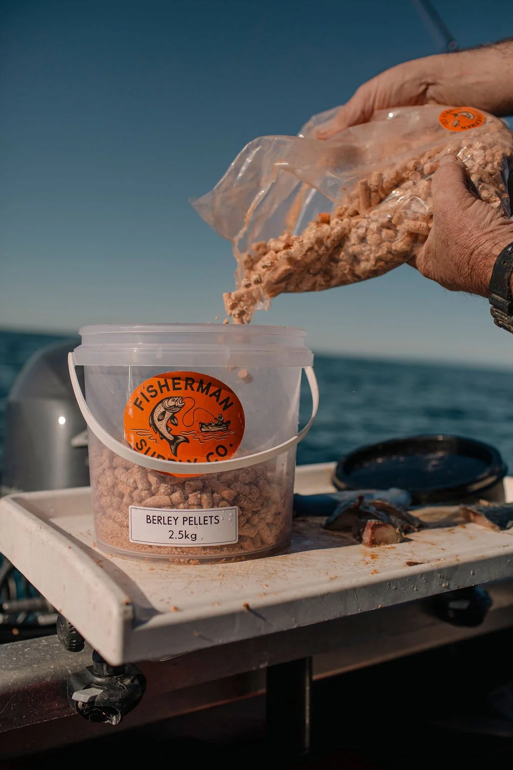 Hands pouring berley pellets from a plastic bag into a container on a boat, with the ocean in the background.