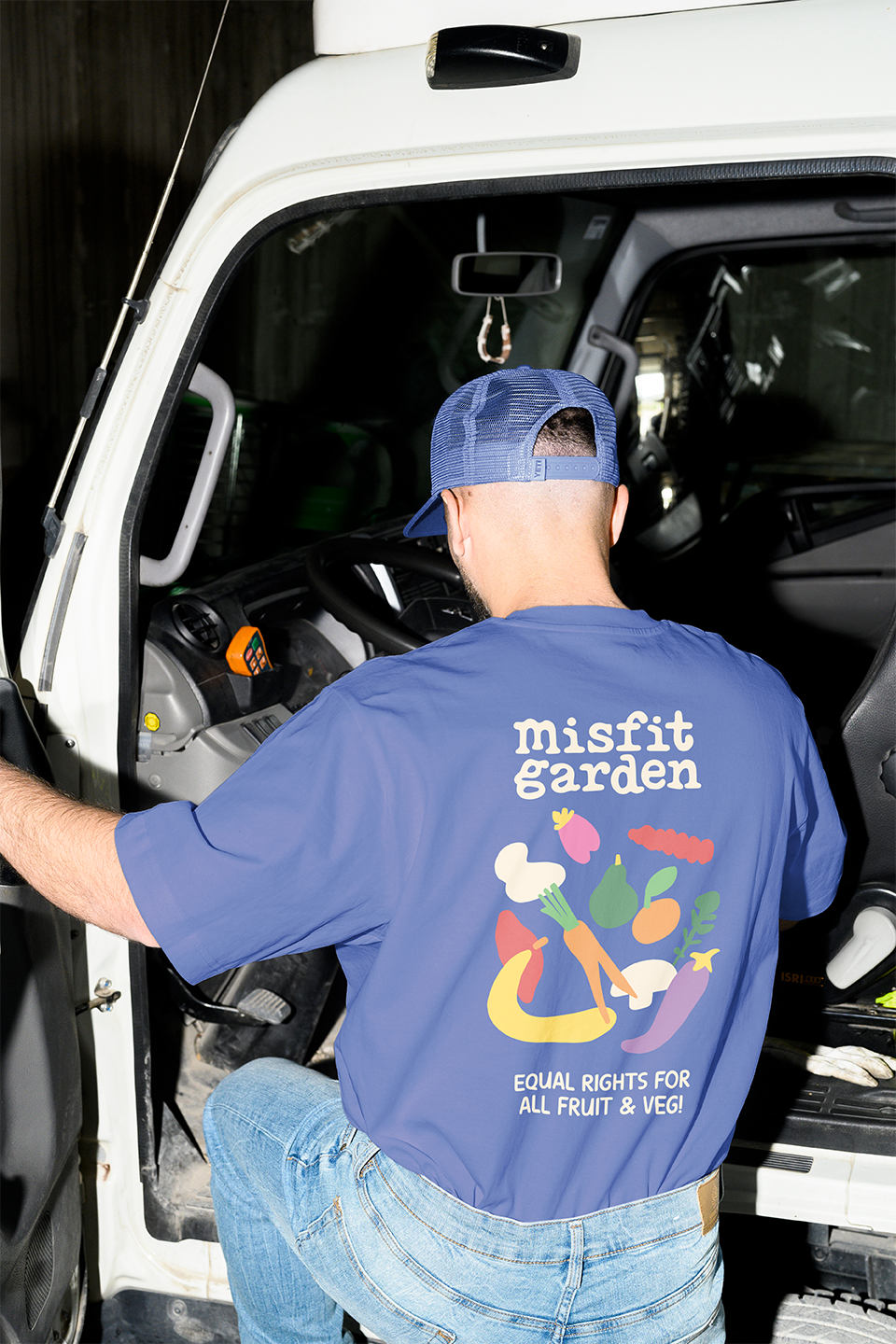 A man wearing a blue cap and t-shirt getting into a white truck, with the back of the shirt displaying a 'Misfit Garden' logo and illustrations of fruits and vegetables.