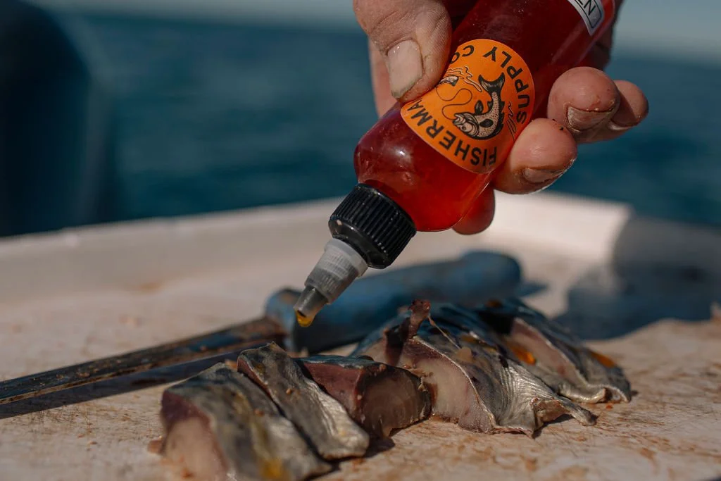 A person holds a bottle of hot sauce over fish fillets on a cutting board.
