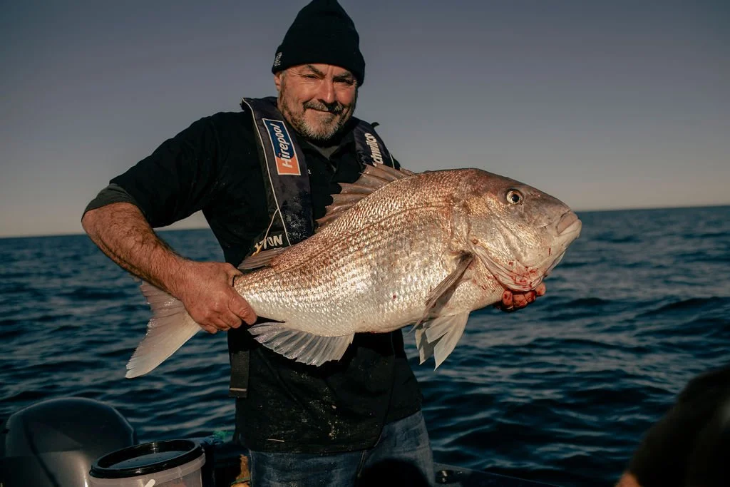 A man in a black beanie and fishing gear holding a large fish on a boat in the ocean during sunset or sunrise.
