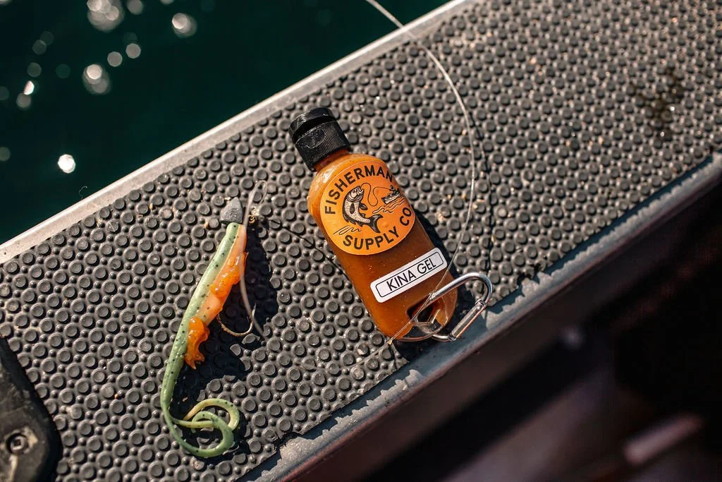 Fishing lure and fishing gel container placed on a boat's textured surface near water.
