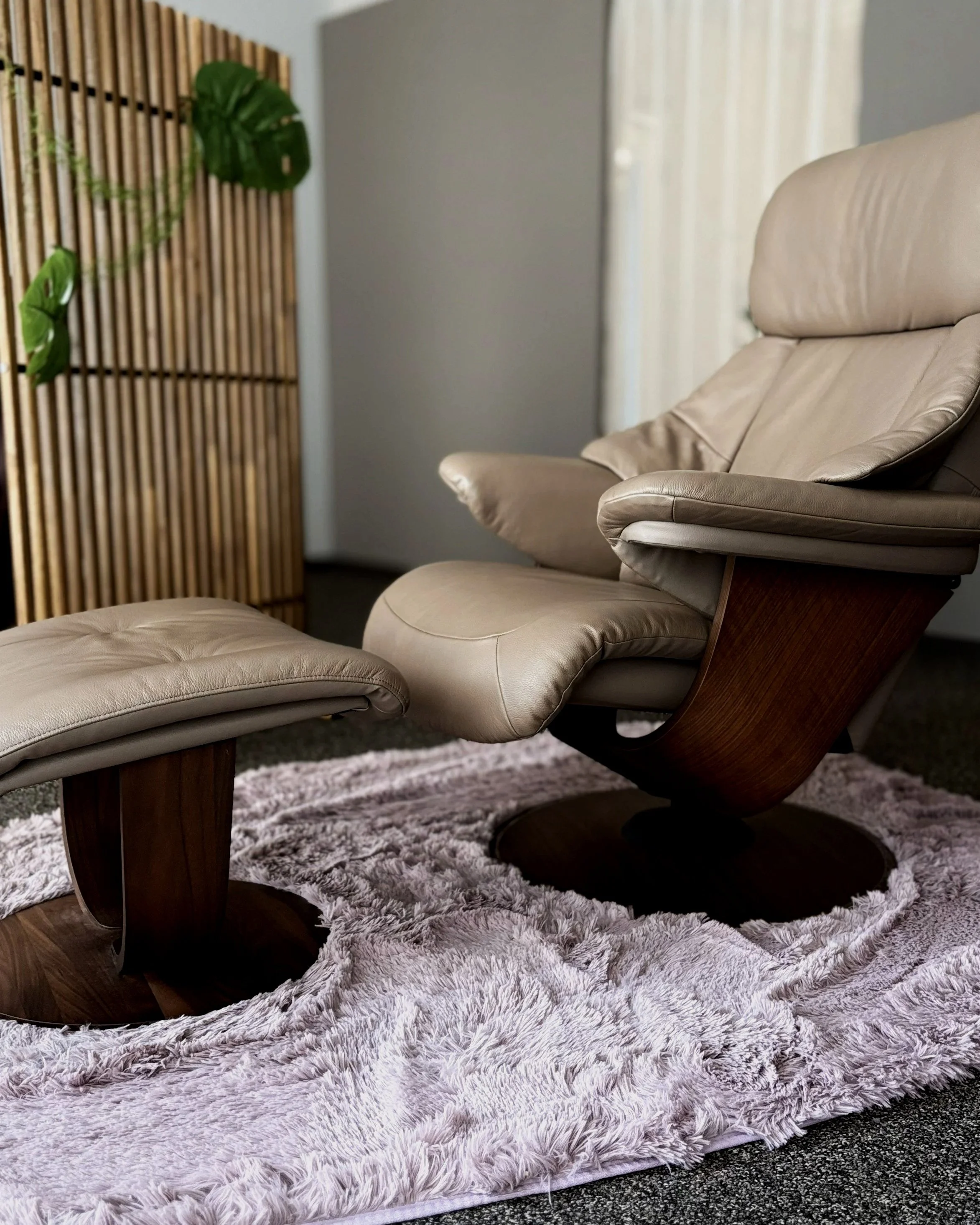 木製のフロアラックの前に置かれた白いレザー製のリクライニングチェアとオットマン。ピンク色の厚手のラグの上に置かれている。