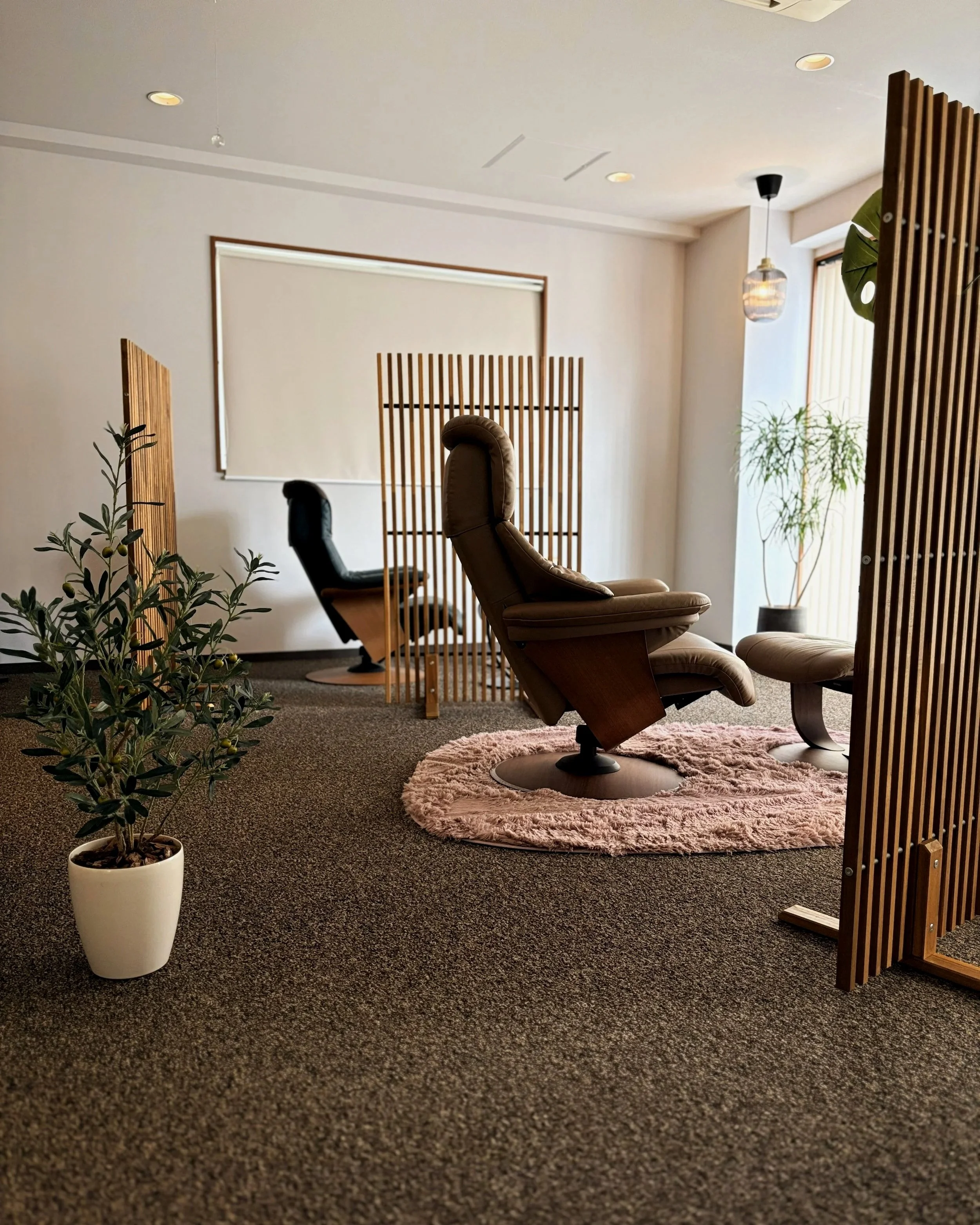 木製の仕切りとリクライニングチェアが置かれたモダンなインテリアのリラックスルーム。窓辺に観葉植物があり、柔らかい照明が灯る空間。