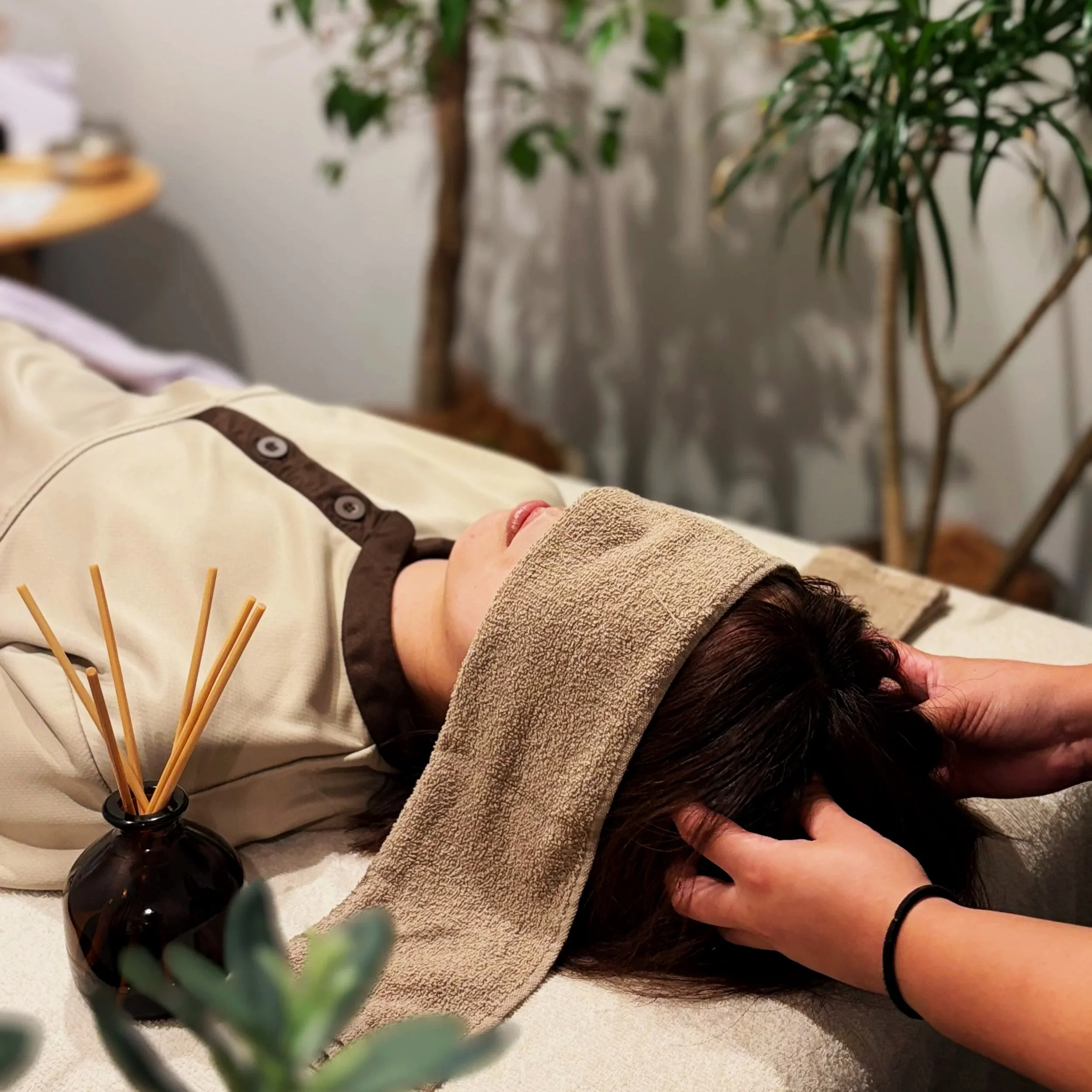 リラックスしている女性が横になり、マッサージ師に首と頭を揉まれている。背後には木や観葉植物があり、リラックスできる環境が整っている