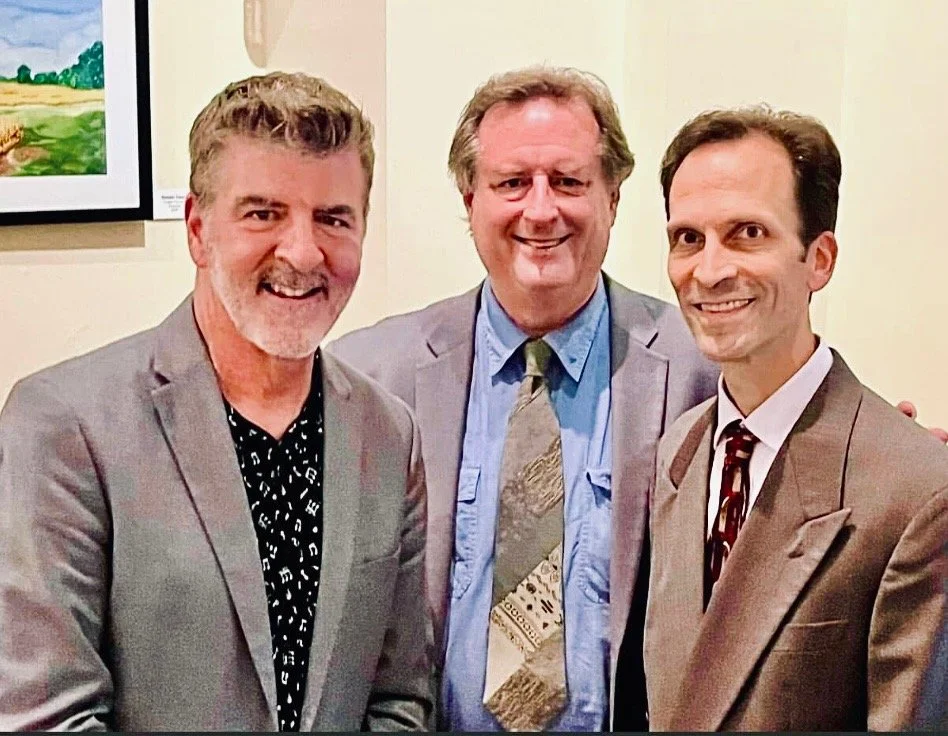 Three men in suits standing together, smiling in front of a black curtain.