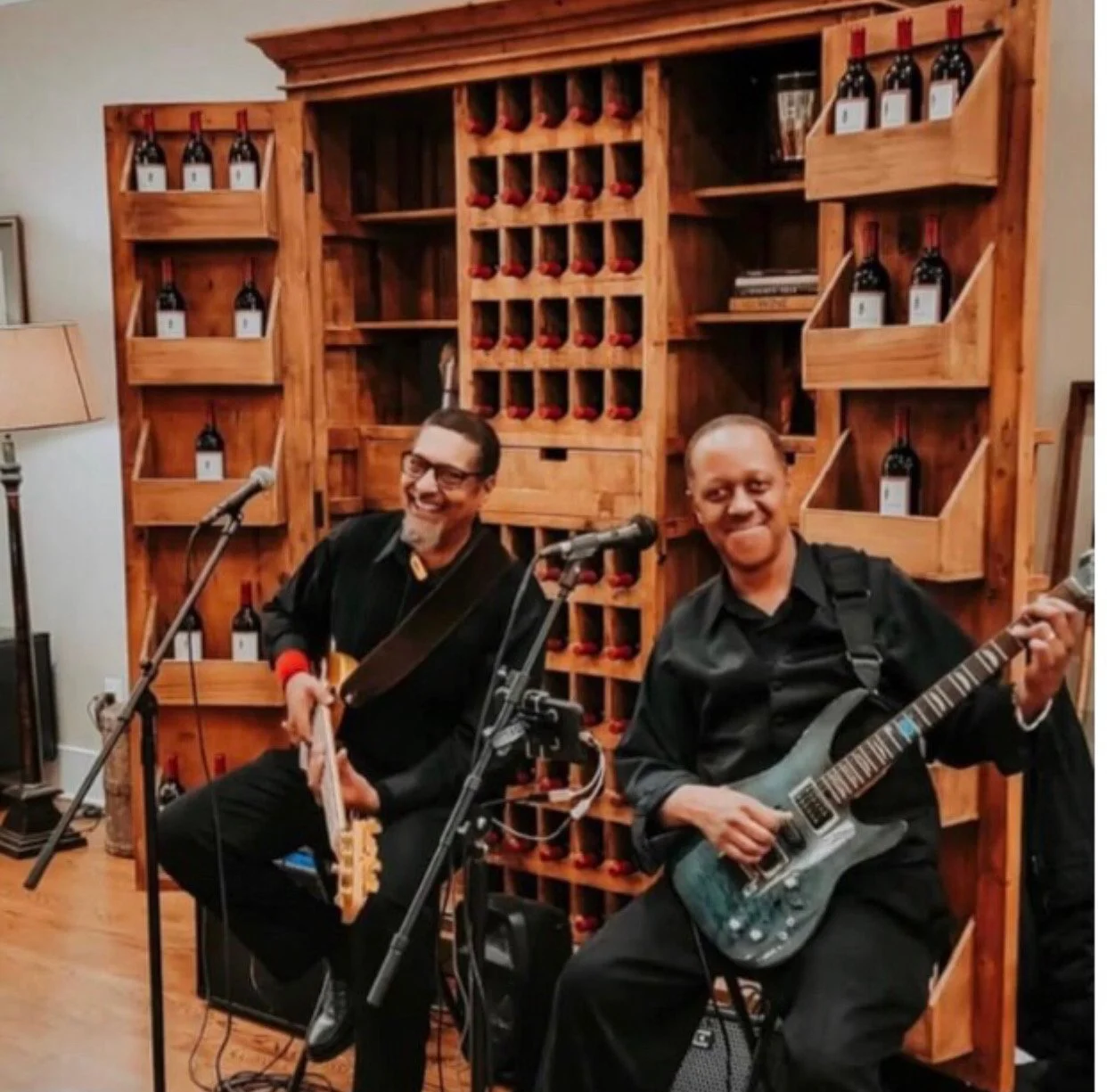 Two men in black shirts holding musical instruments and smiling, standing in front of a dark curtain.