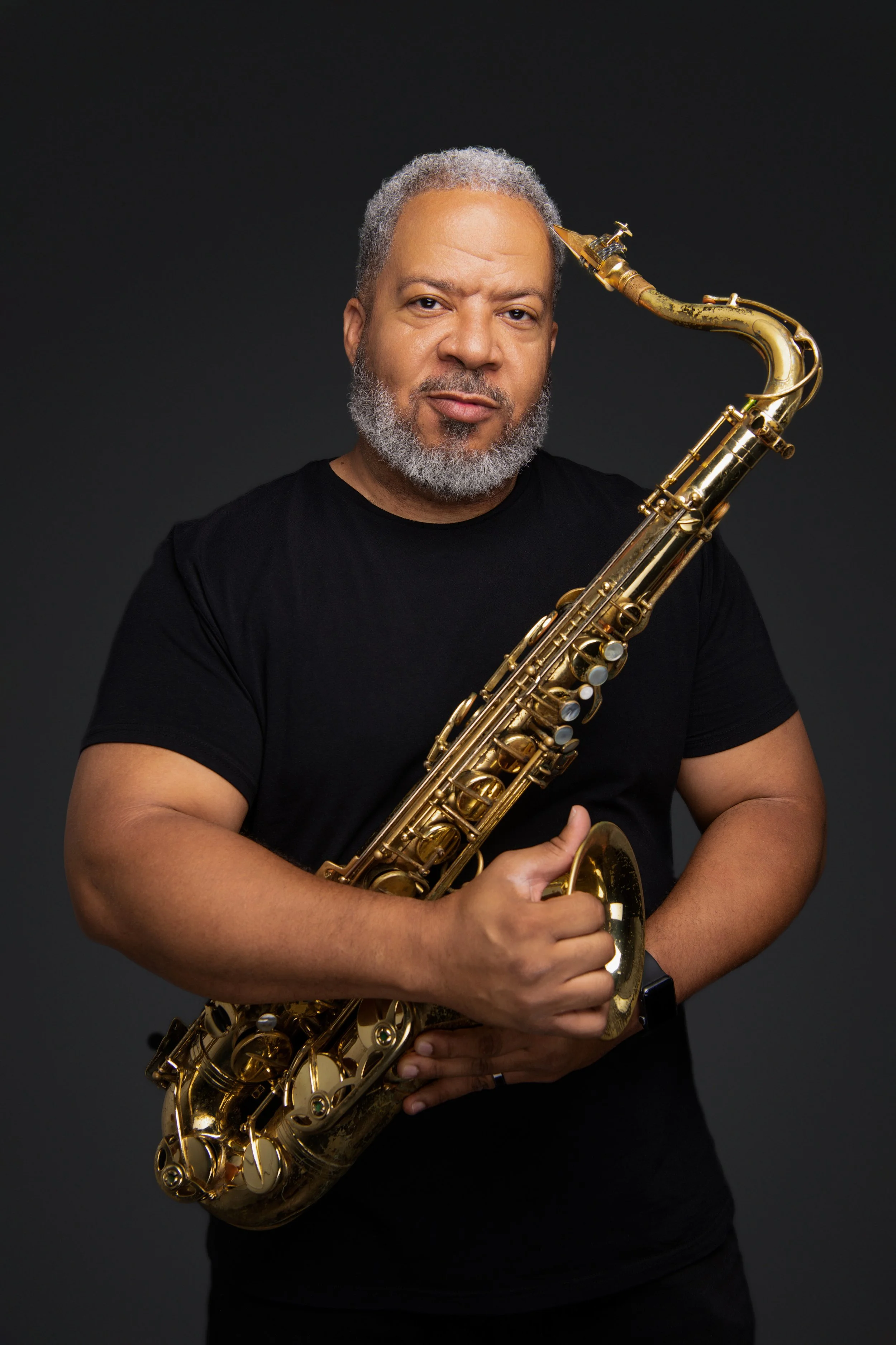 A man with gray hair and a beard holding a gold saxophone against a dark background.