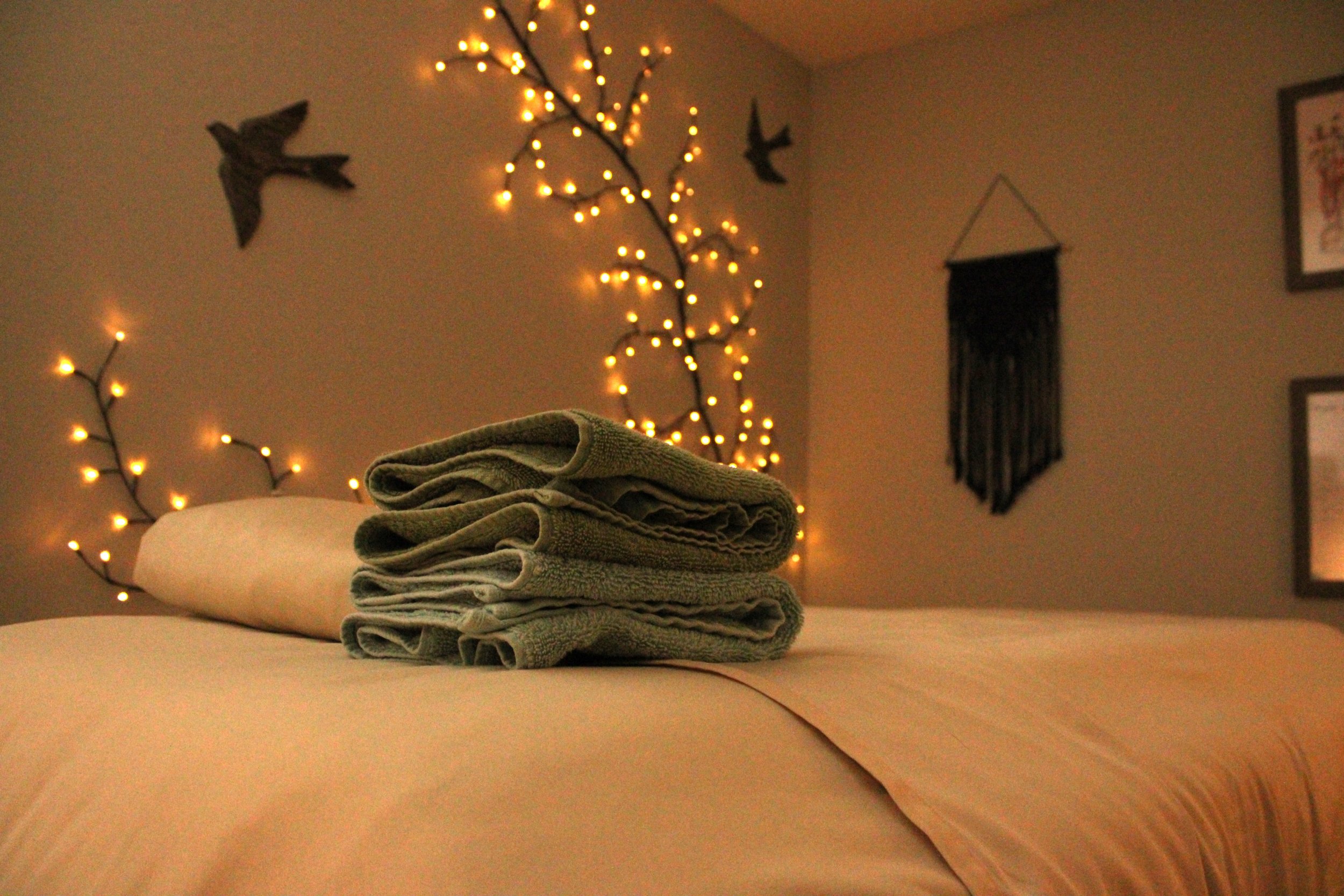 Warm amber lit room with massage table, freshly folded towels and linens sit on the head of the bed. A branch of fairy lights sits on the background wall with flying sparrow silhouettes dotting the wall.