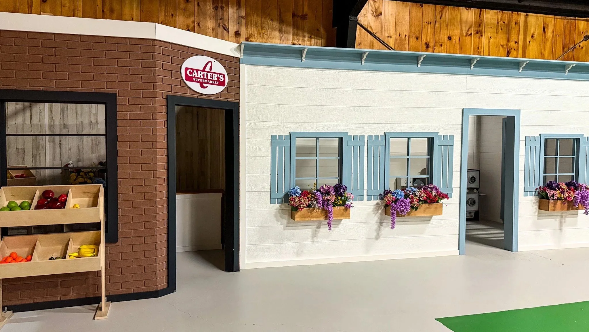Miniature model of a supermarket storefront with a sign that reads Carter's Supermarket, featuring a white building with light blue window shutters and flower boxes, a brick wall on the left side, and a fruit stand with plastic fruit in front.